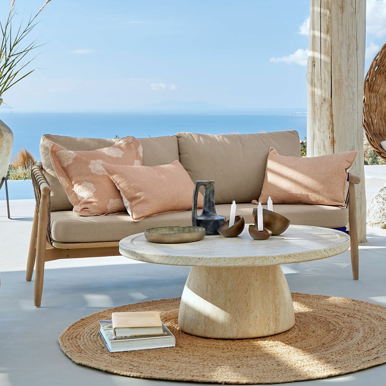 Divano FSC®-100% Thornwick Divano beige con cuscini e tavolo rotondo in pietra su terrazza con vista mare