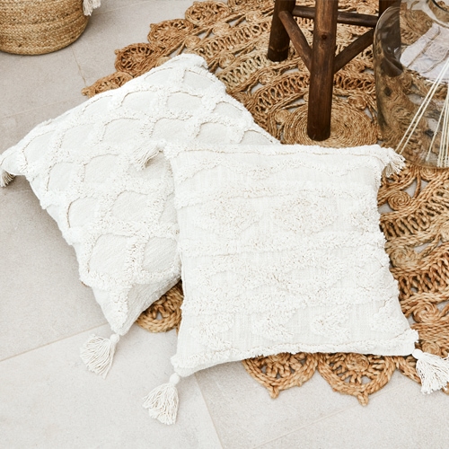 Due cuscini decorativi crema in cotone con nappine agli angoli su tappeto intrecciato