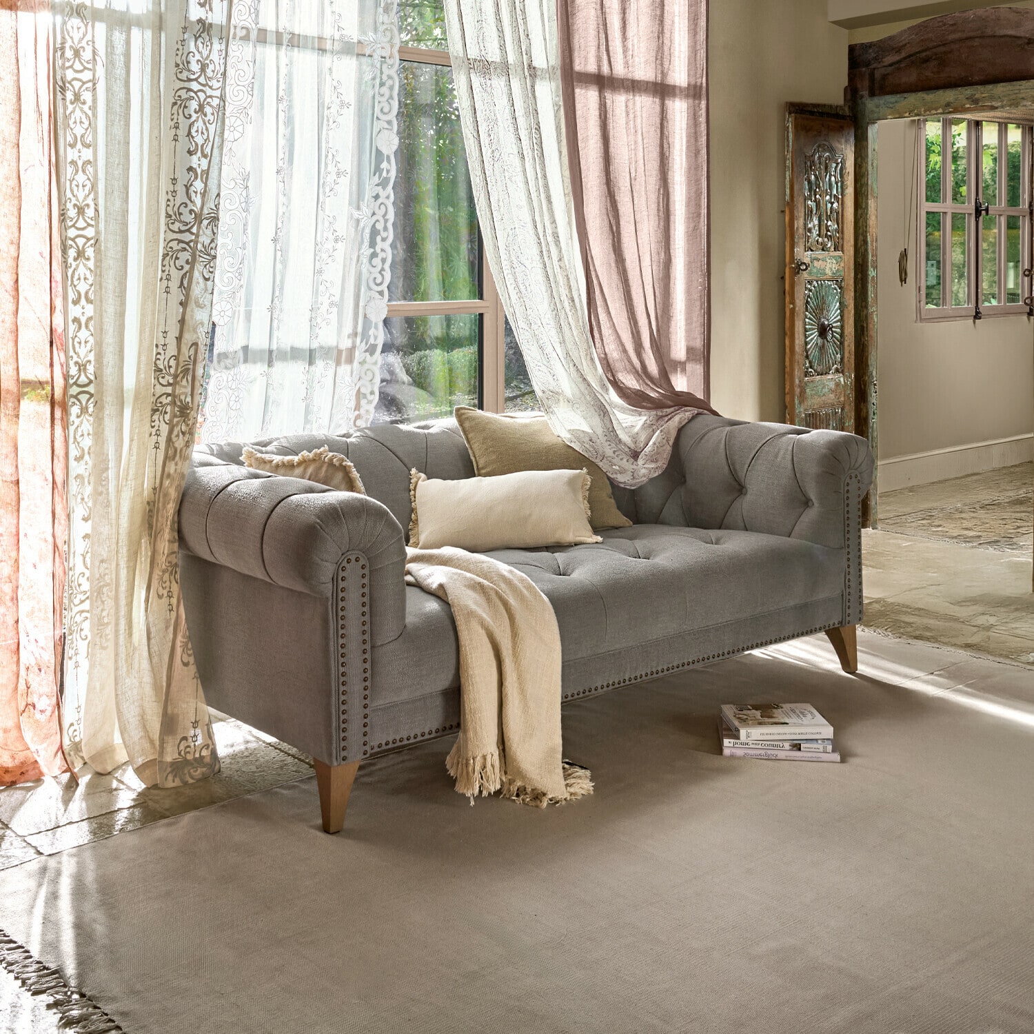 Divano grigio imbottito con gambe in legno e cuscini beige in soggiorno luminoso con tende