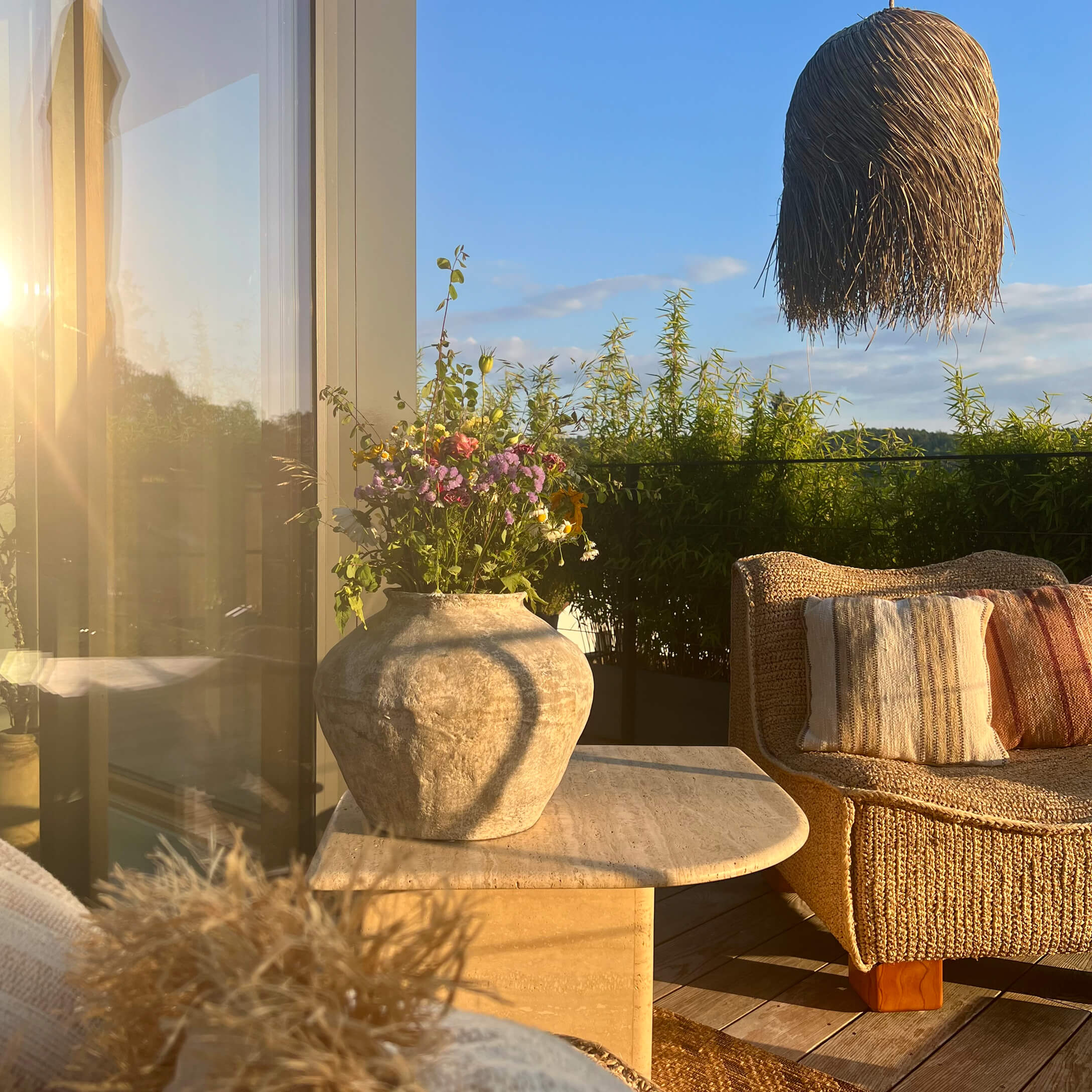 Grande vaso in pietra su tavolo accanto a poltrona intrecciata con fiori e piante in terrazza