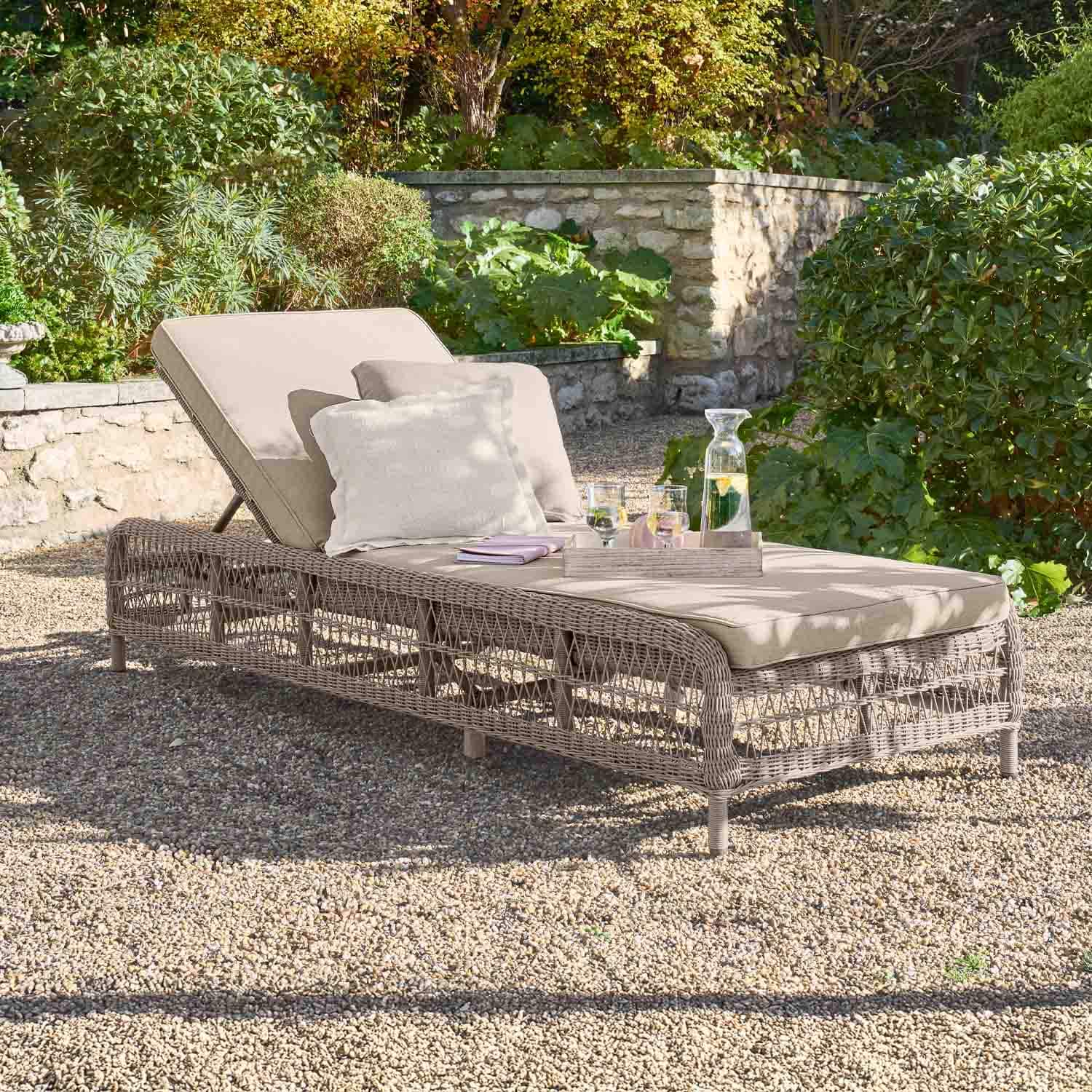 Lettino da giardino beige con cuscino e struttura in vimini su ghiaia in giardino verde