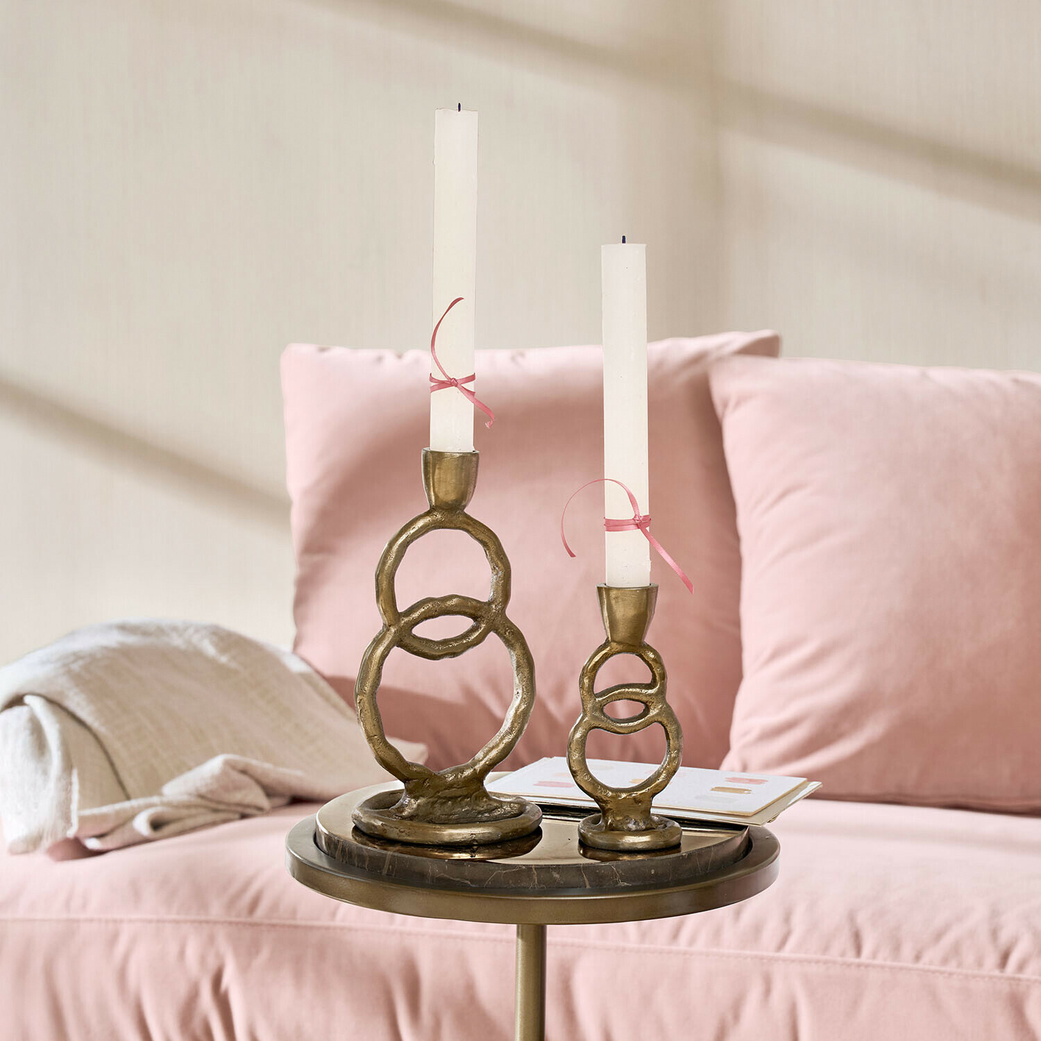 Coppia di portacandele Corvane Set di due portacandele in ottone con candele bianche su un tavolino accanto a un divano rosa