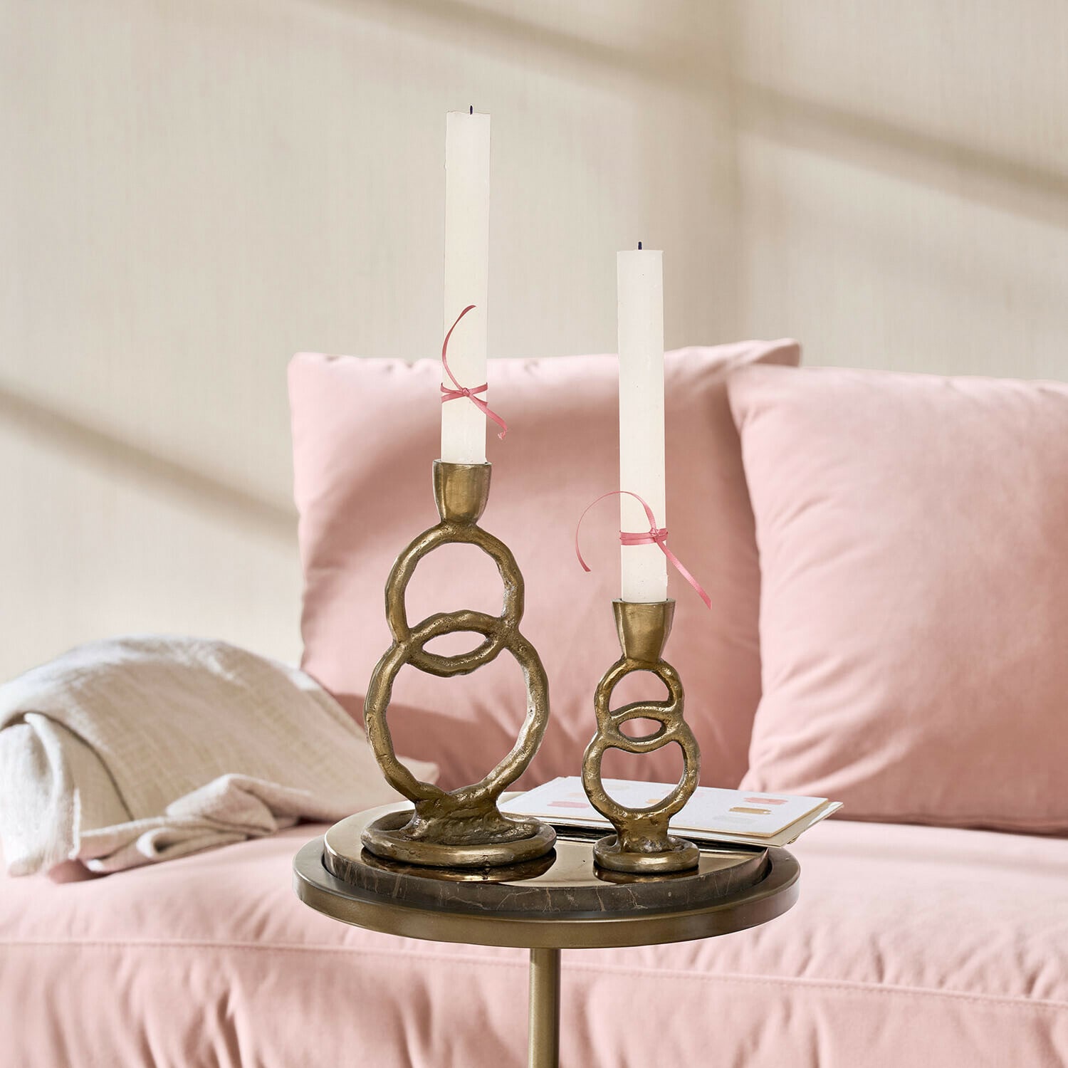 Coppia di portacandele Corvane Set di due portacandele in ottone con candele bianche su un tavolino accanto a un divano rosa