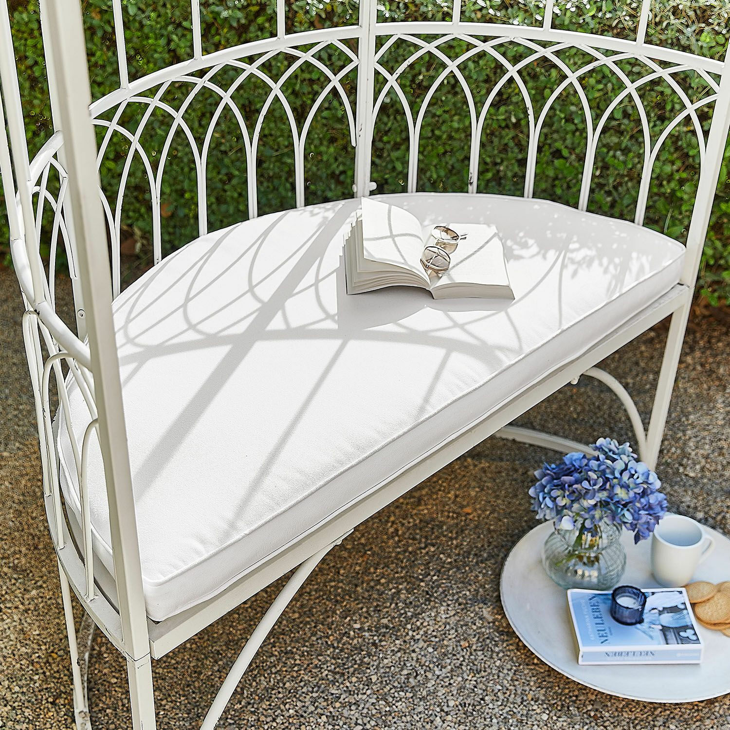 Cuscino da seduta Drivesto Panca da giardino in metallo bianco con cuscino bianco, libro aperto, fiori blu e tazza sul patio