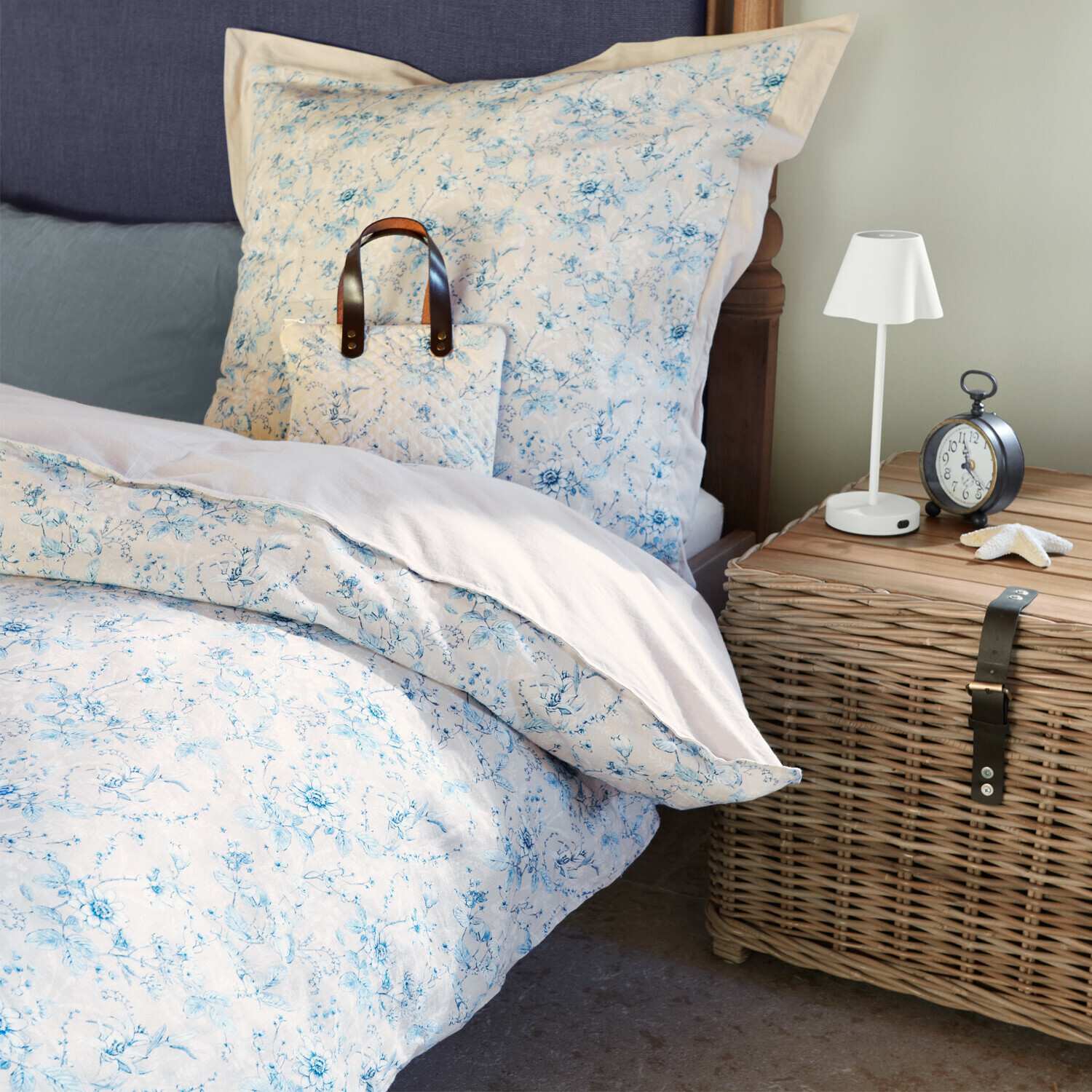 Biancheria da letto in cotone con fiori azzurri chiari su letto accanto a comodino in vimini e lampada