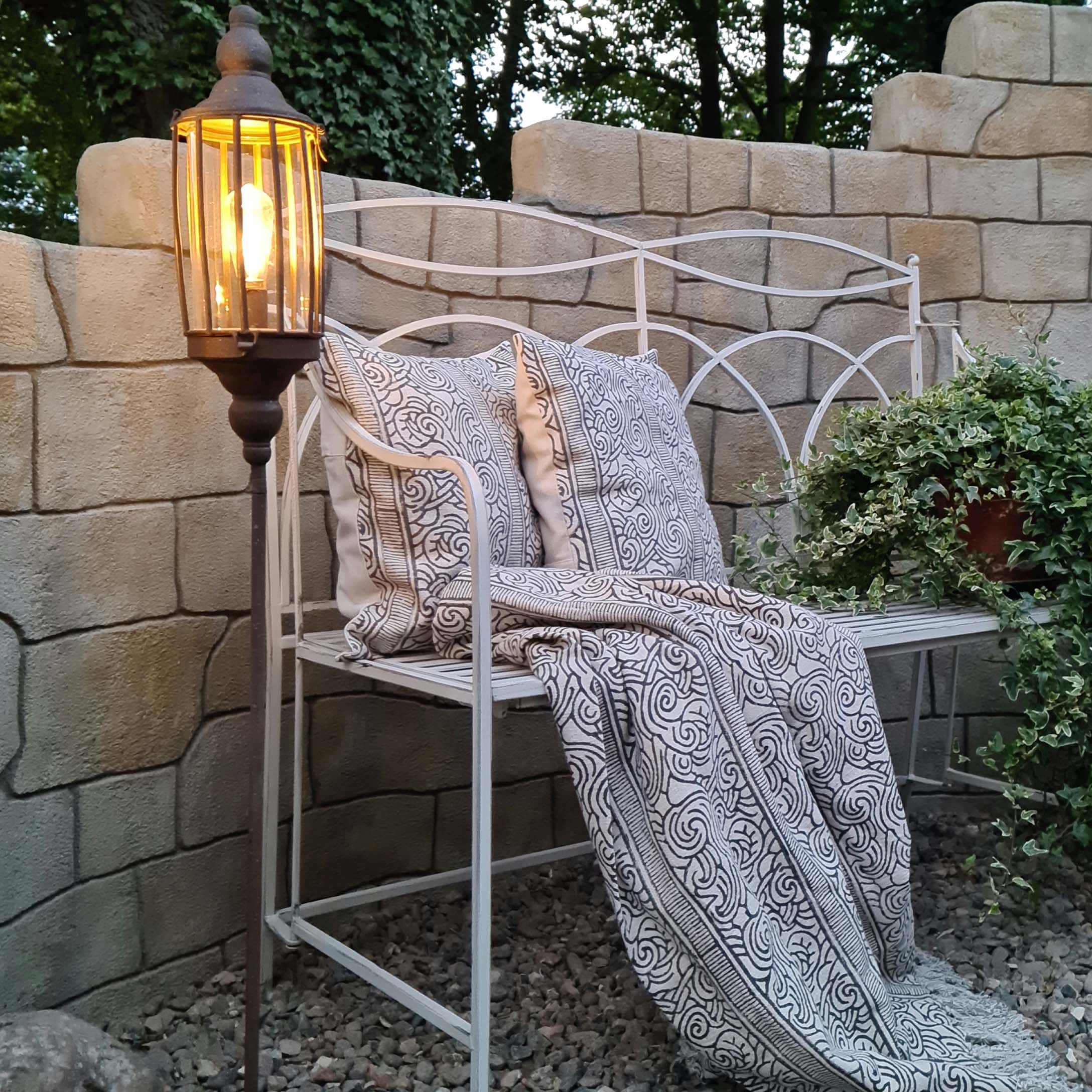 Panchina da giardino in metallo bianco con cuscini e coperta a motivi bianco e nero, lampada da terra accesa