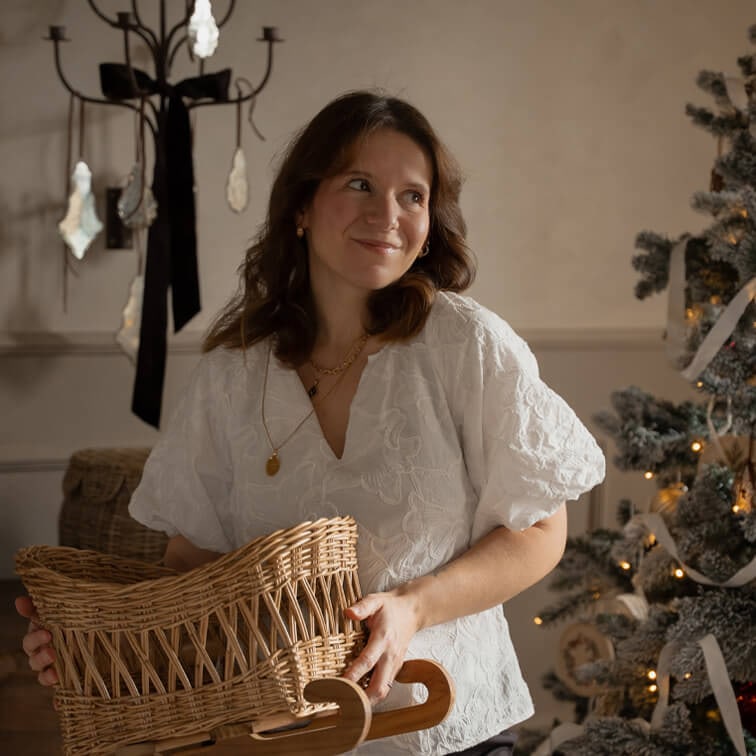Donna con camicetta bianca ricamata che tiene un cesto di vimini su slitta di legno vicino all'albero di Natale decorato