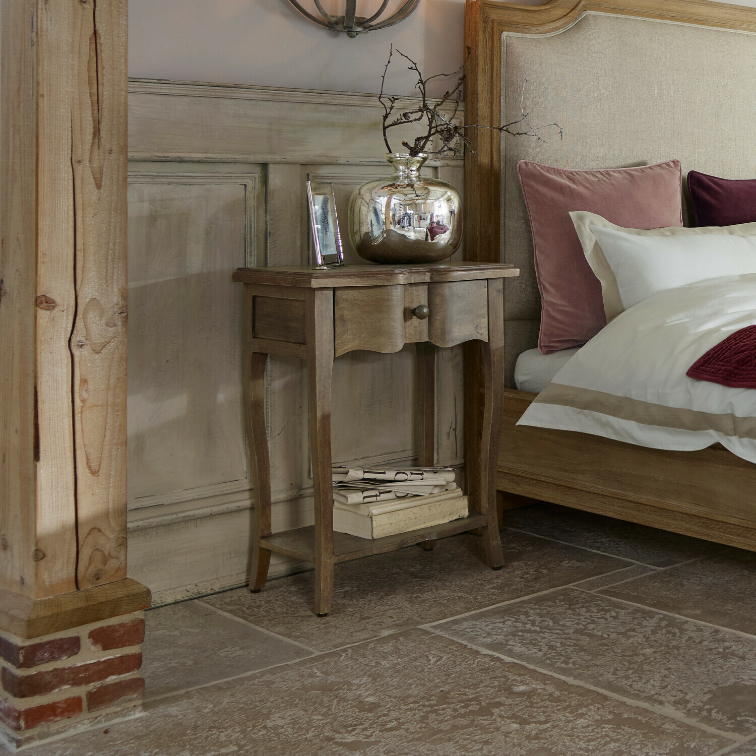 Tavolino di complemento Finstock Comodino in legno con cassetto e ripiano, decorato con vaso argentato e libri accanto a letto con cuscini rosa