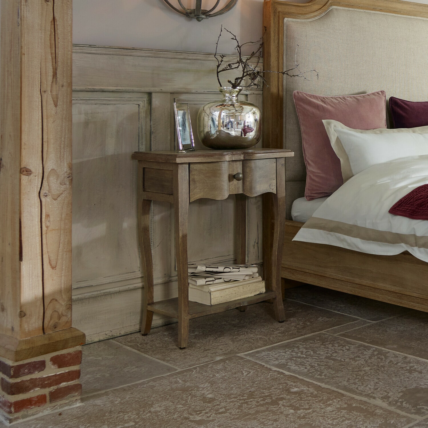 Tavolino di complemento Finstock Comodino in legno con cassetto e ripiano, decorato con vaso argentato e libri accanto a letto con cuscini rosa