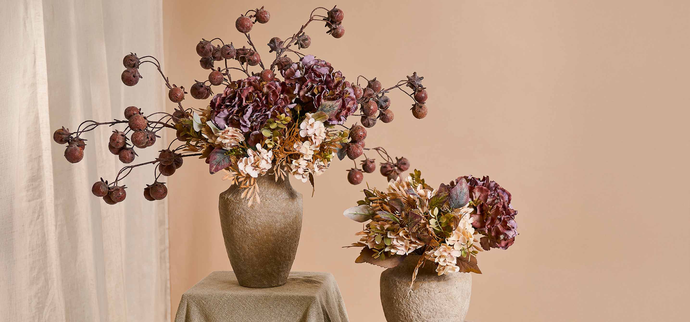 Due vasi decorativi in pietra beige con mazzi di fiori artificiali e frutti dai toni caldi