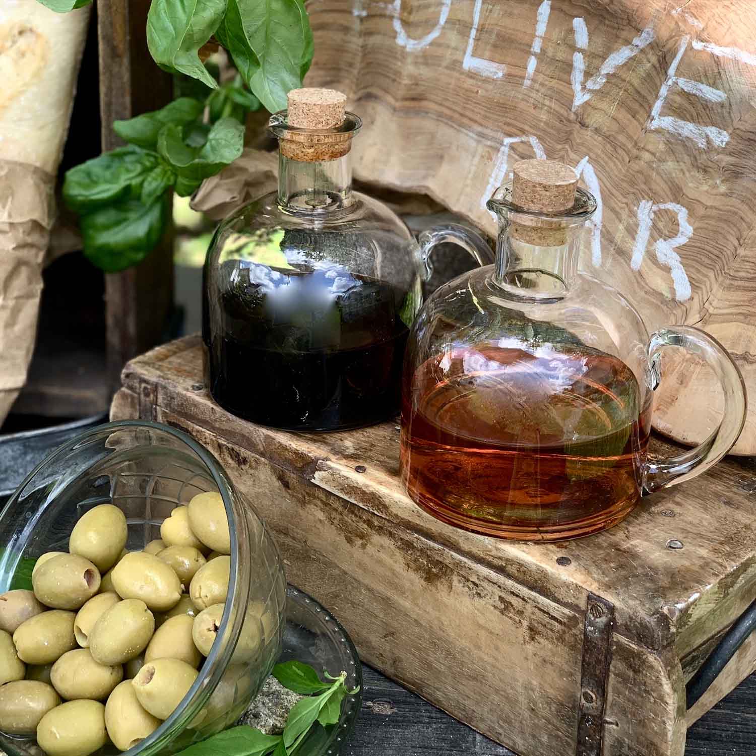 Bottiglie di vetro con tappi in sughero con olio e aceto su cassa di legno accanto a olive verdi e foglie di basilico