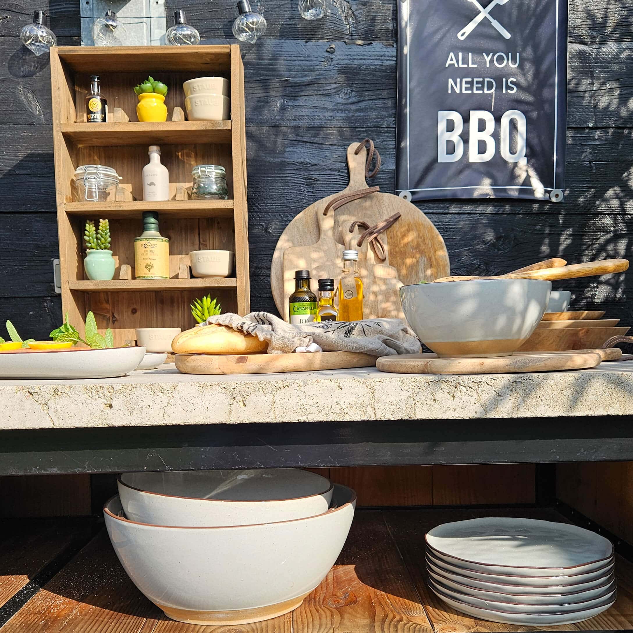 Set di ciotole e piatti in ceramica beige con taglieri in legno e bottiglie di olio su tavolo da barbecue