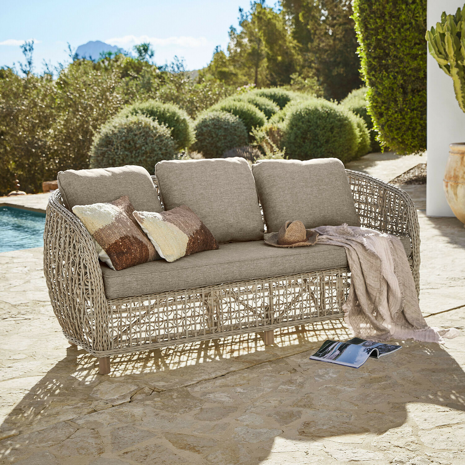 Divano da esterno beige con cuscini e struttura in rattan intrecciato su terrazza in pietra vicino alla piscina