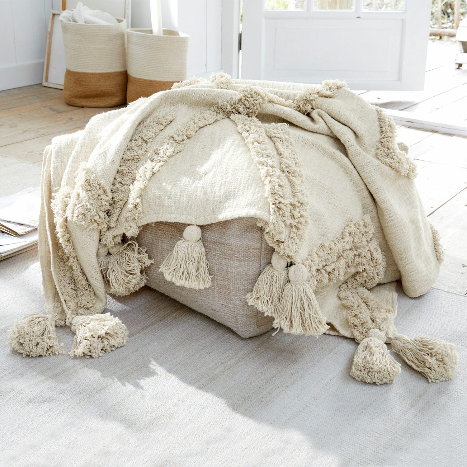 Coperta Linares Coperta beige in cotone con nappine su pouf chiaro in ambiente luminoso naturale