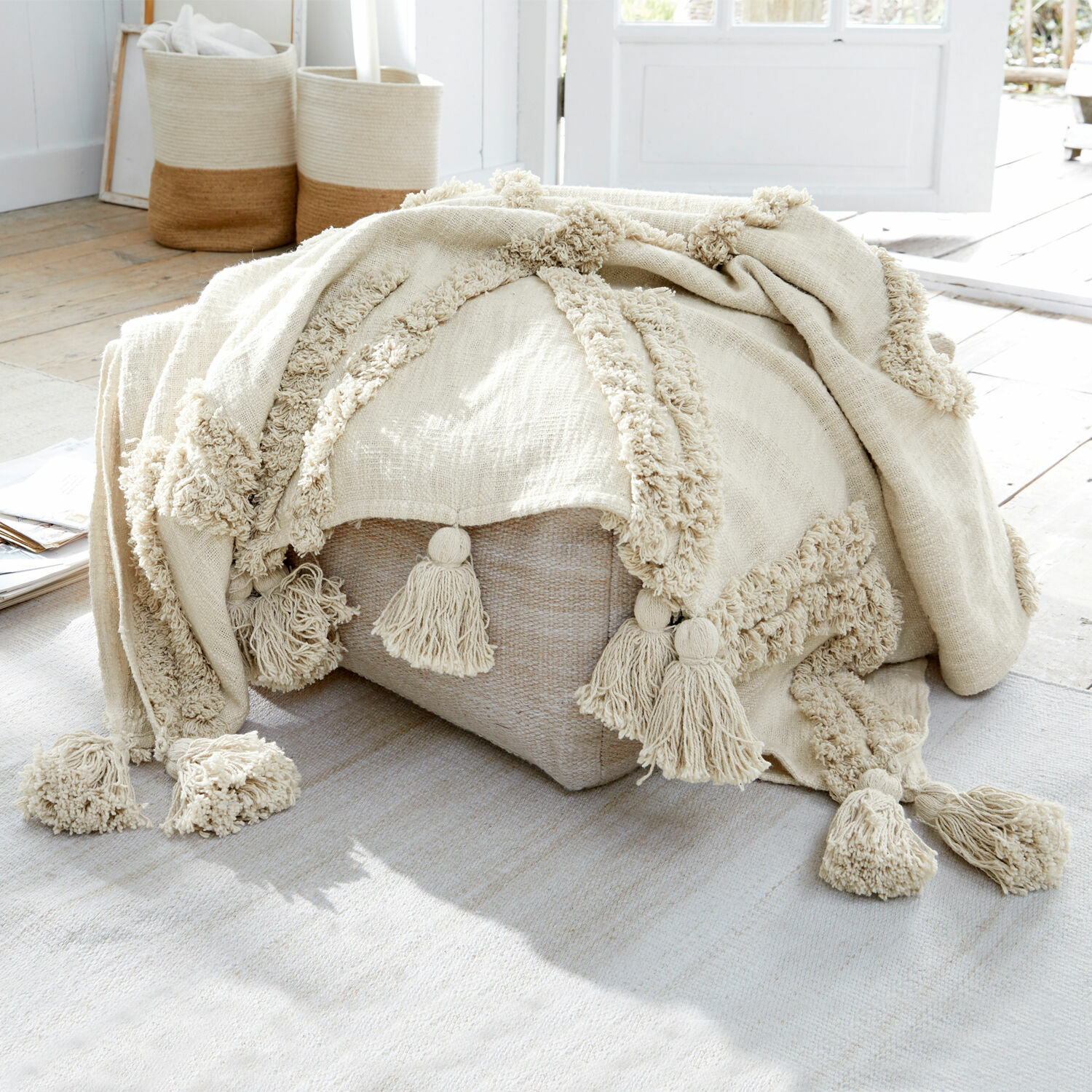 Coperta Linares Coperta beige in cotone con nappine su pouf chiaro in ambiente luminoso naturale