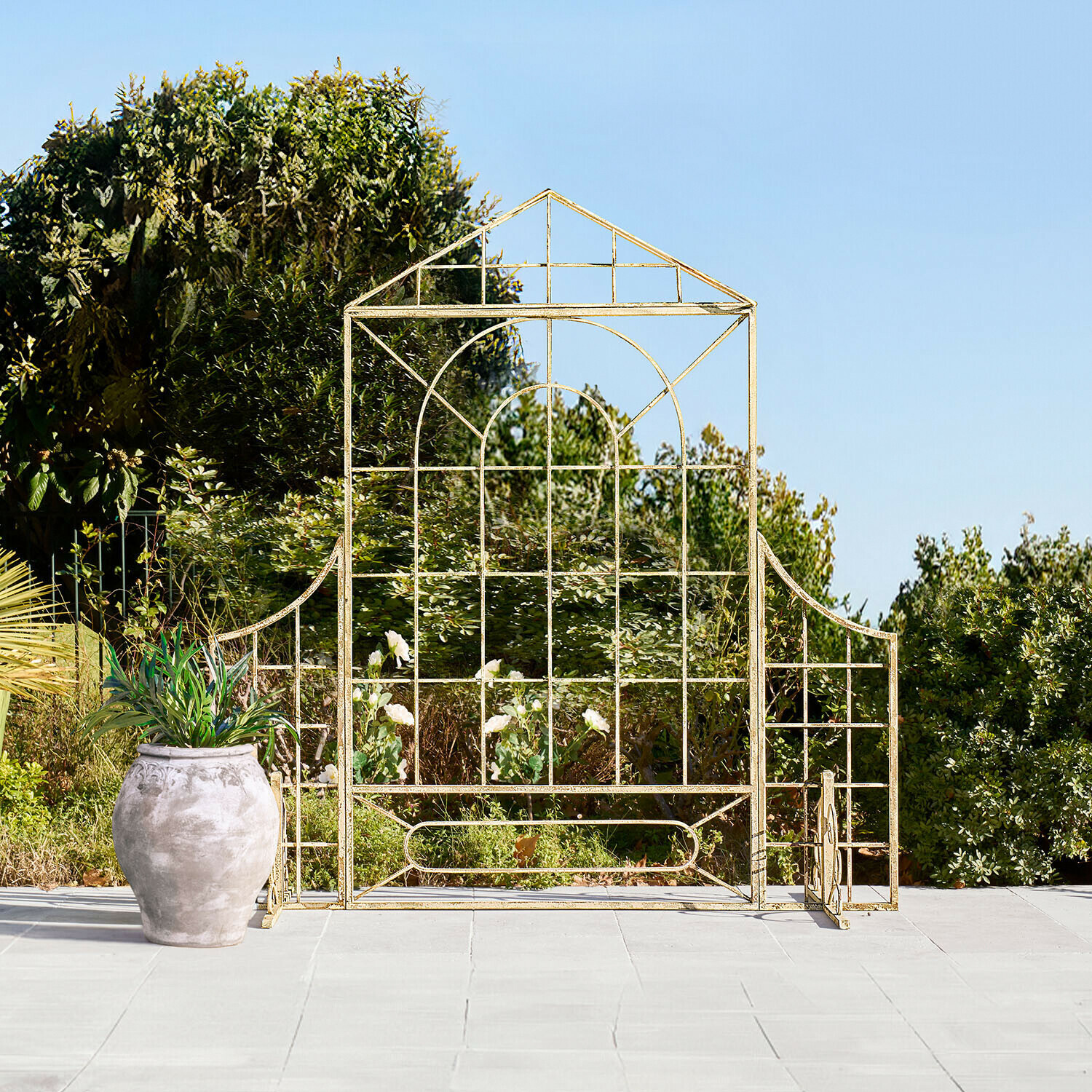 Griglia da giardino in metallo dorato a forma di casa su terrazza con vaso e vegetazione sullo sfondo