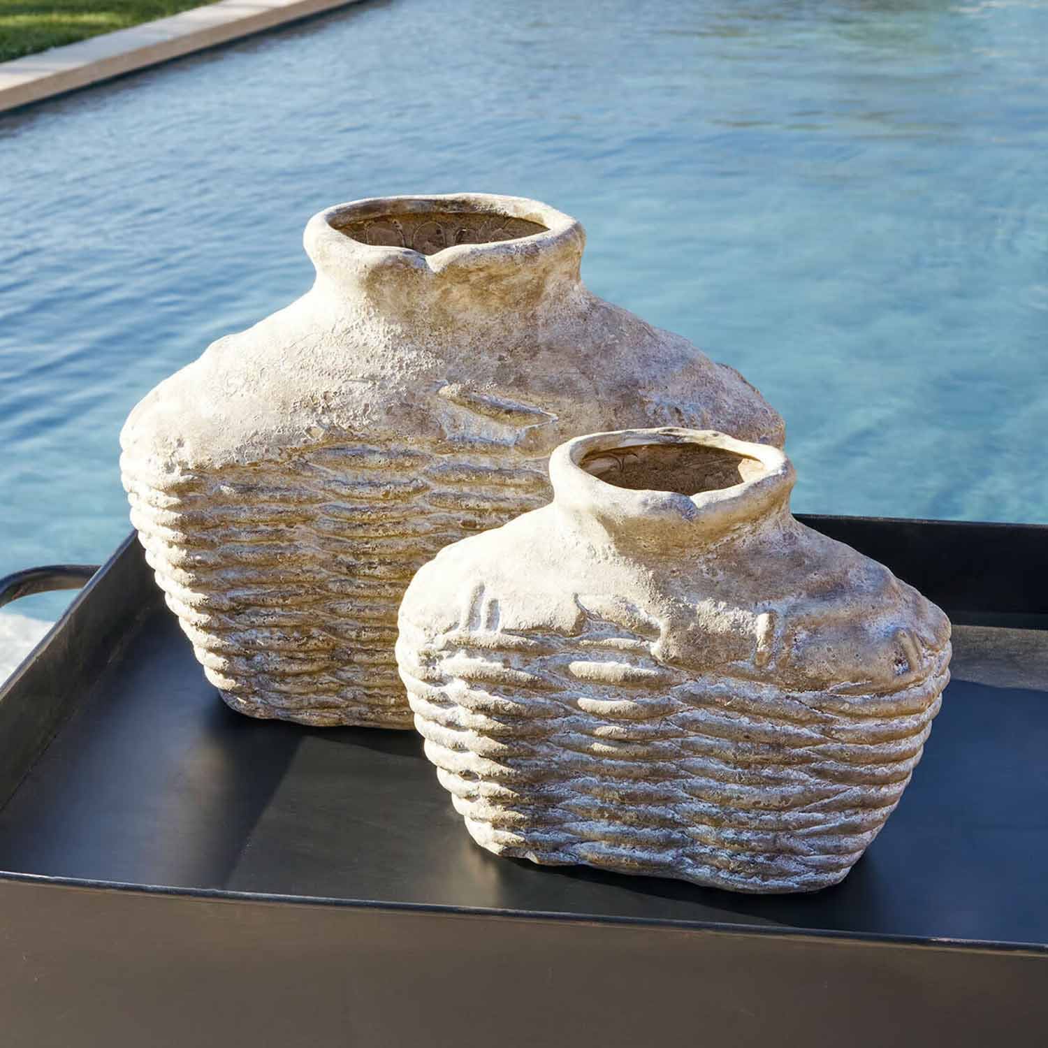 Due vasi rustici in pietra beige su un vassoio nero vicino a una piscina