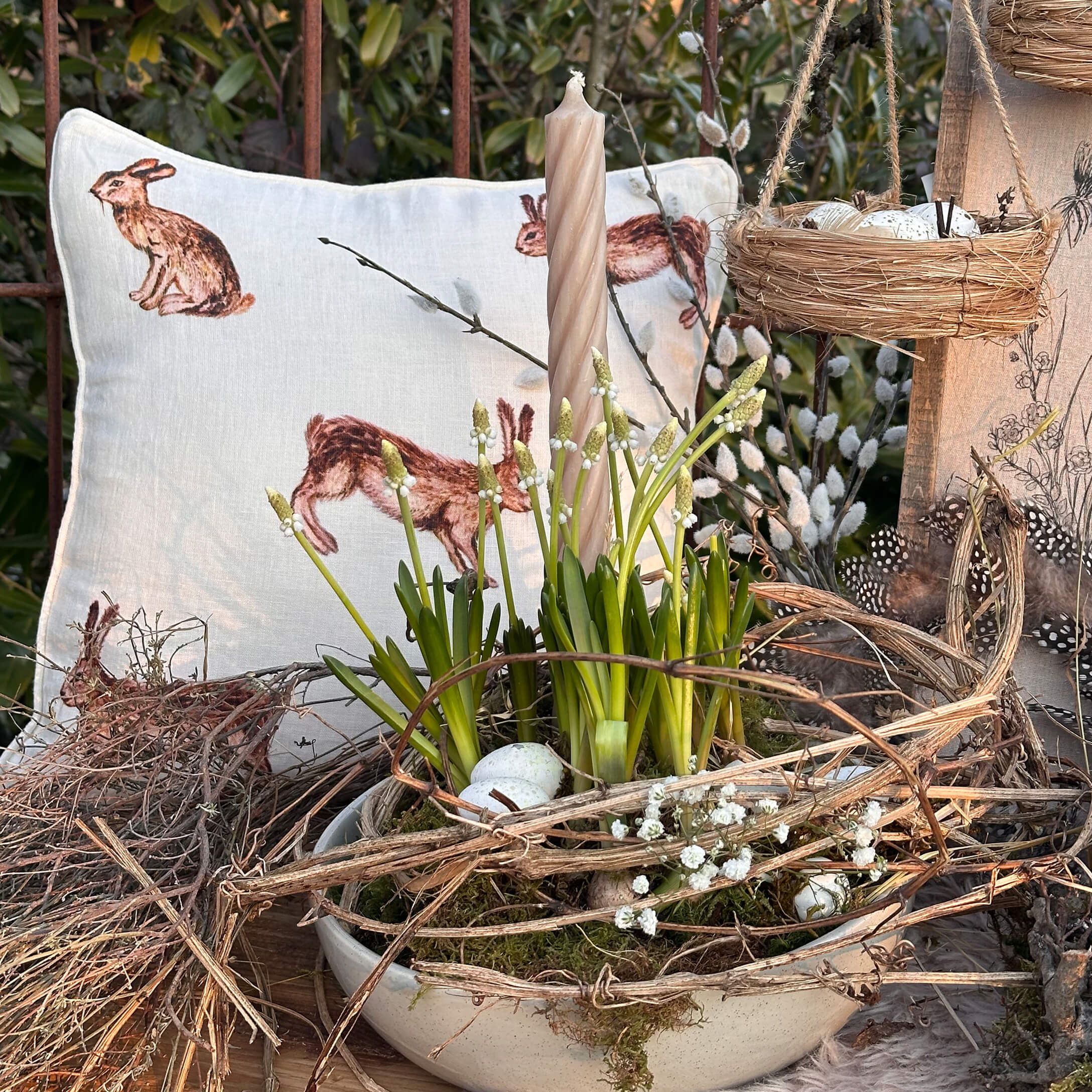 Decorazione pasquale con candela bianca, fiori verdi e cuscino con motivo coniglio su tavolo in legno