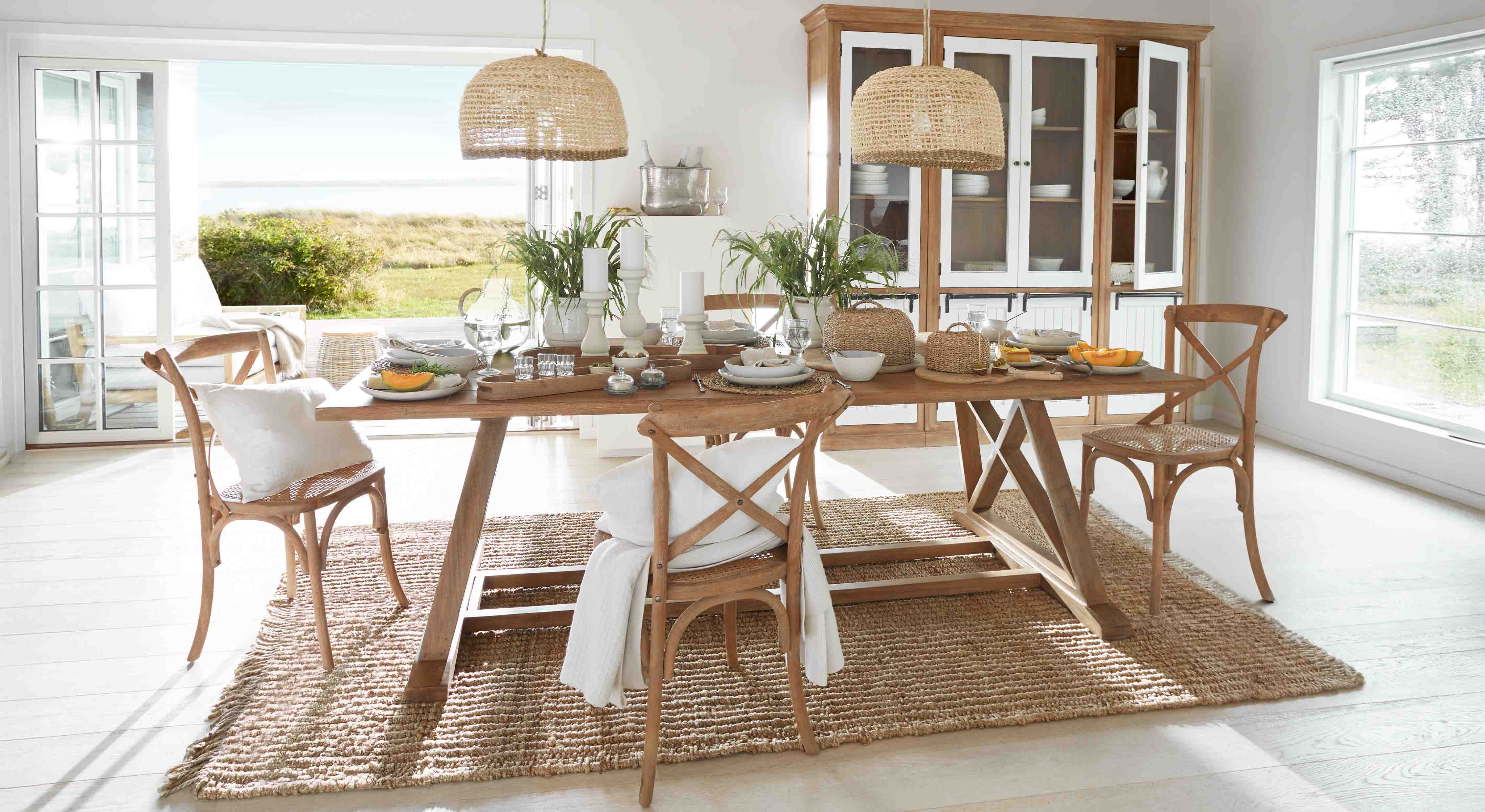 Sala da pranzo con tavolo in legno e sedie in rattan su tappeto naturale, decorazioni vegetali e lampade in vimini
