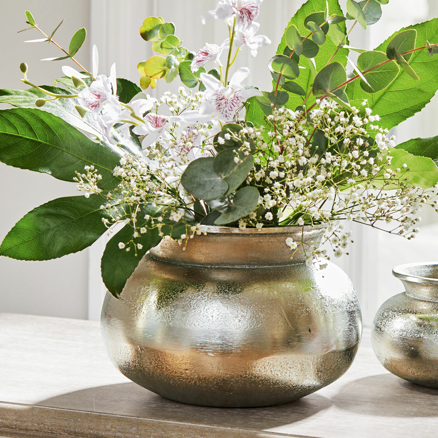 Vaso Hadrien Vaso in metallo argentato con fiori bianchi e foglie verdi su tavolo in legno, decorazione d'interni