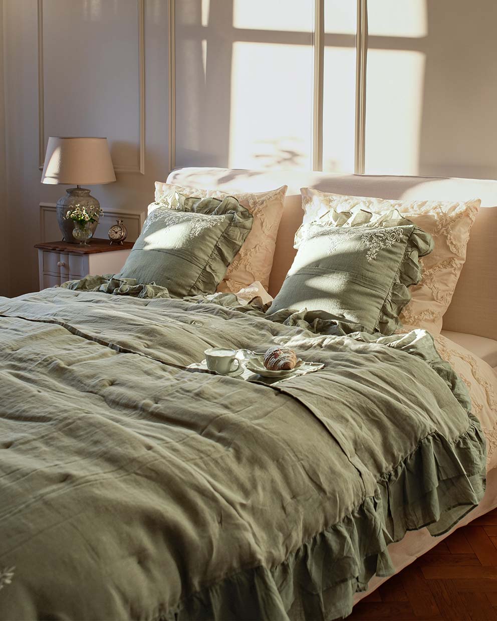 Biancheria da letto verde in lino con volant e cuscini ricamati in camera luminosa con lampada e orologio