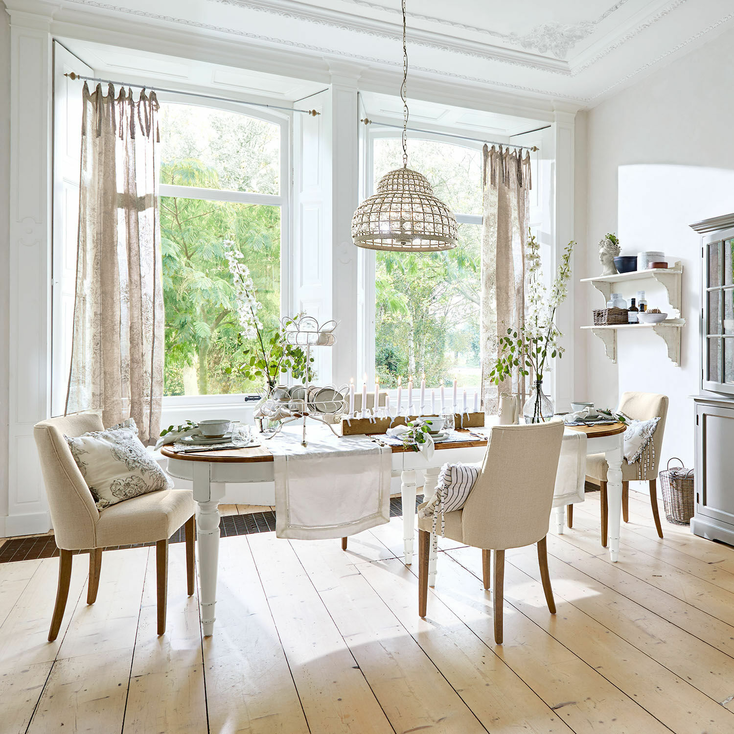 Tavolo da pranzo Pinecrest Sala da pranzo con tavolo in legno bianco e sedie beige su pavimento in legno chiaro, decorazioni e lampada a sospensione in vimini