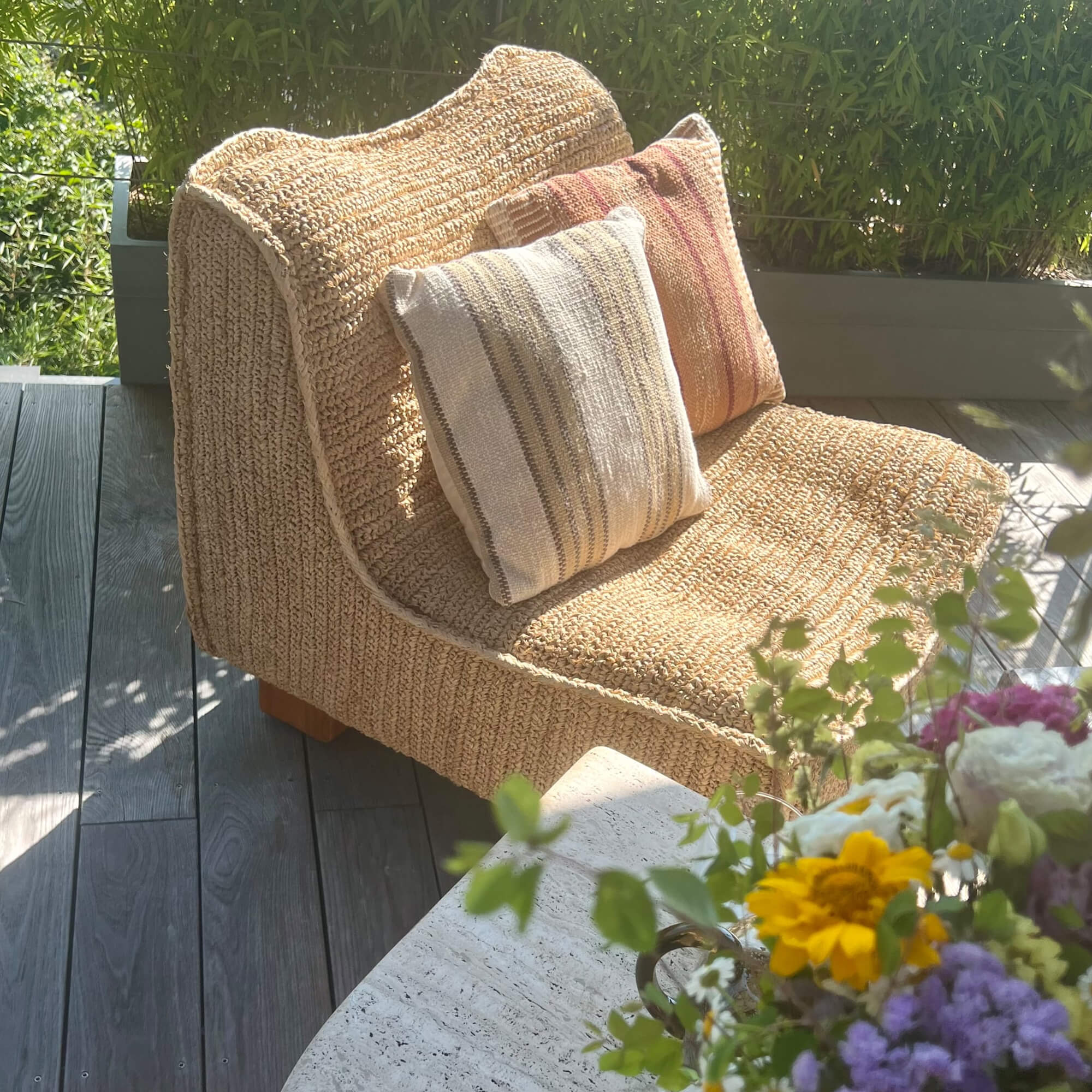 Poltrona beige in materiale intrecciato con due cuscini decorativi su terrazza fiorita
