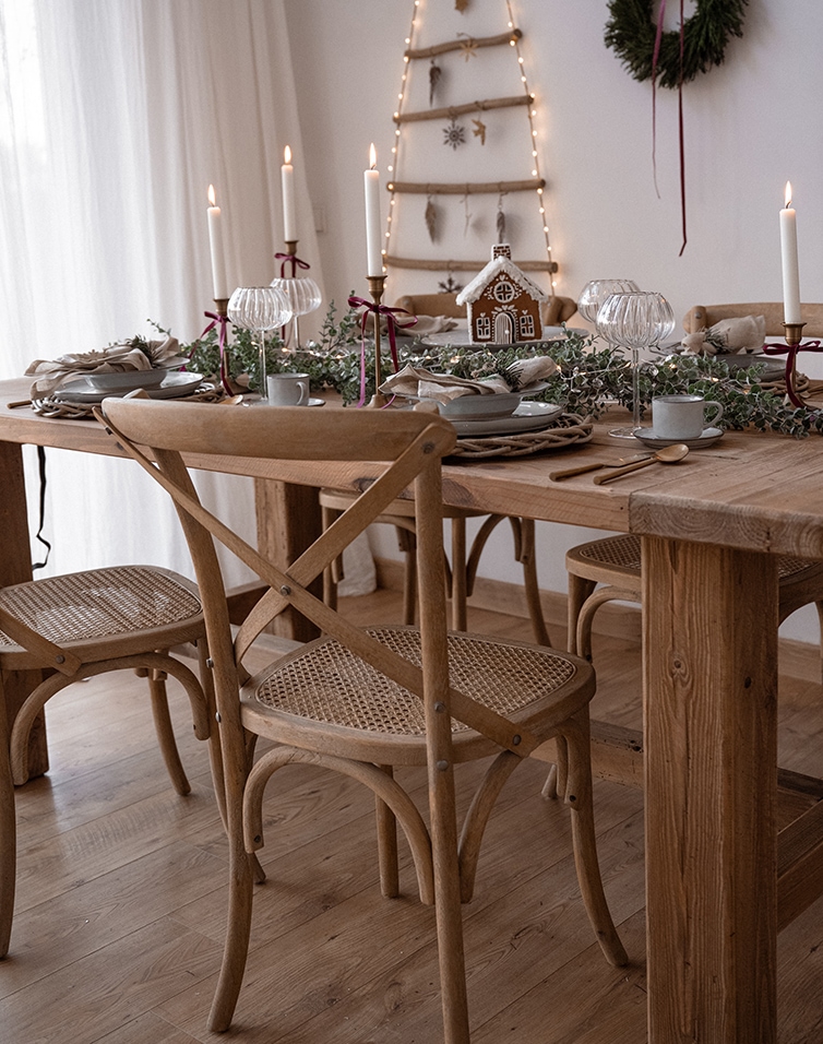 Tavola di Natale in legno con decorazioni naturali, candele e sedie rustiche in ambiente luminoso