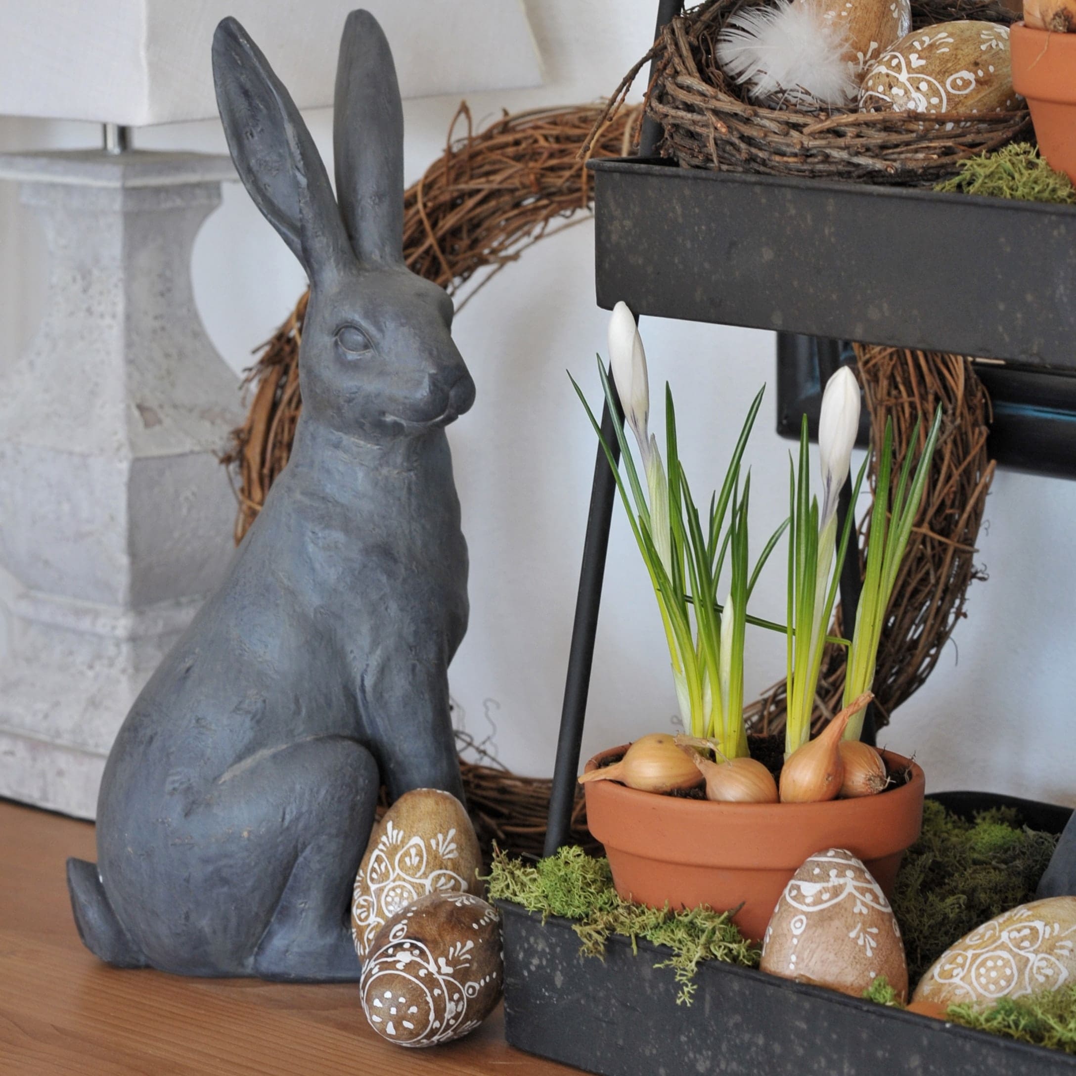 Coniglio decorativo grigio in ceramica accanto a vaso con fiori bianchi e uova pasquali decorate