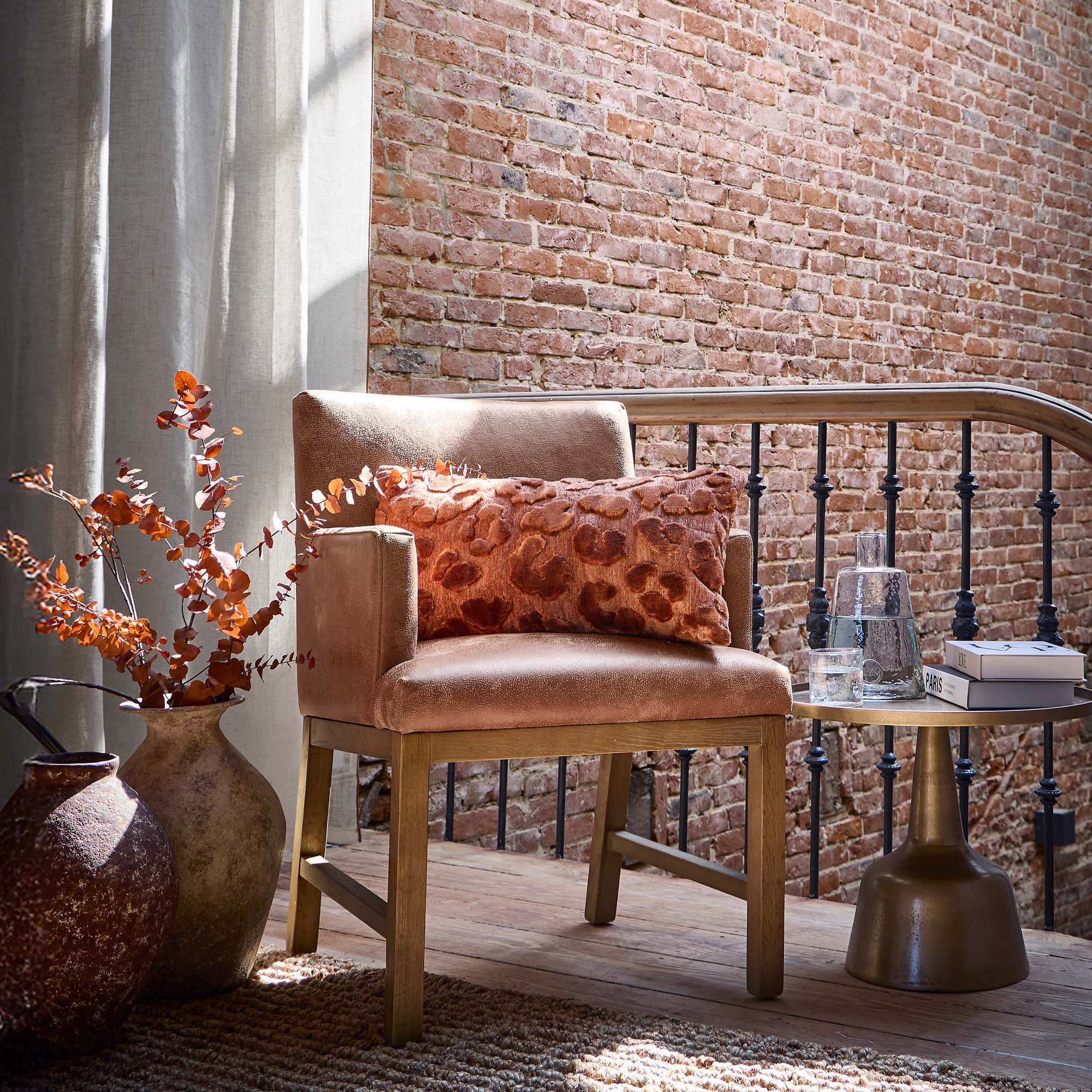 Poltrona beige con base in legno e cuscino arancione davanti a muro di mattoni e tende