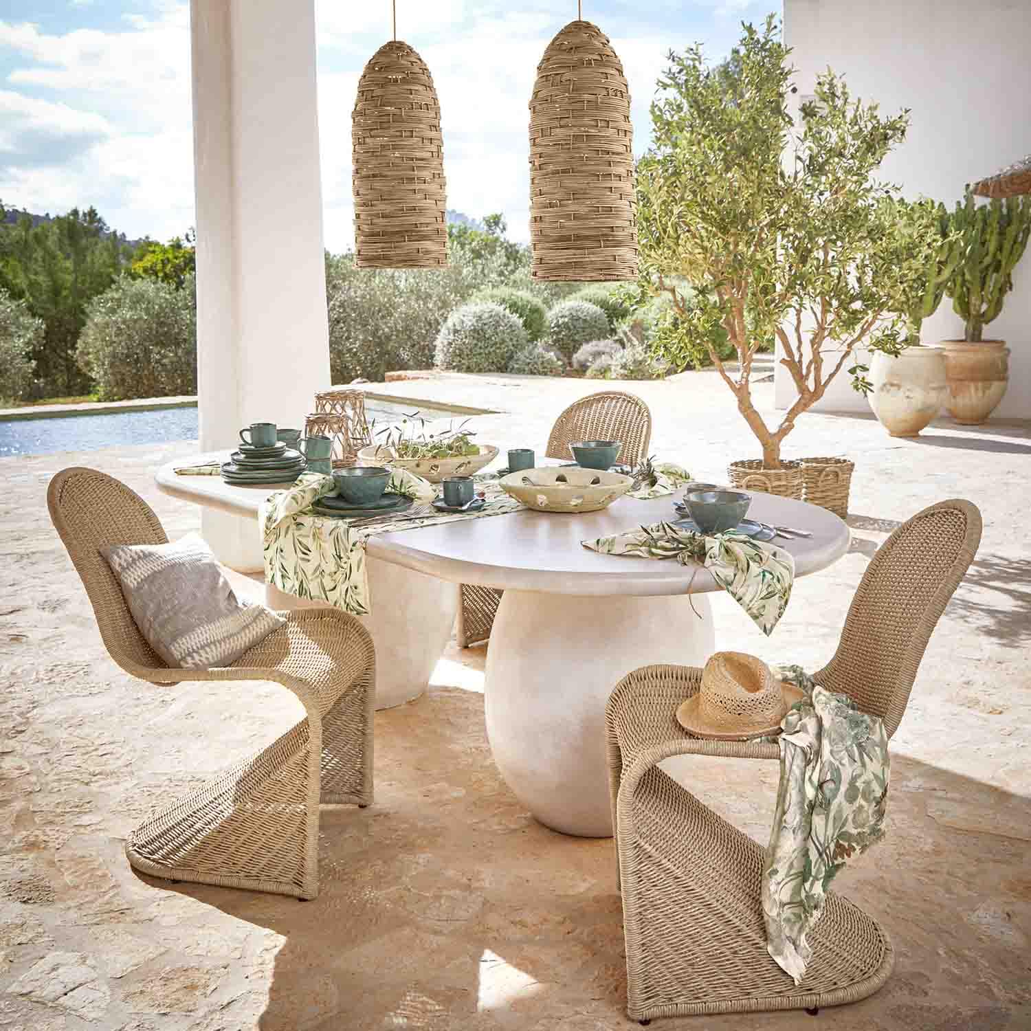Tavolo ovale bianco in pietra con sedie in rattan e lampade sospese naturali in giardino soleggiato