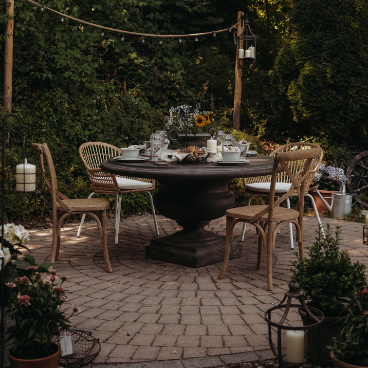 Tavolo rotondo da giardino in legno scuro con sedie in vimini su patio lastricato decorato con candele e fiori