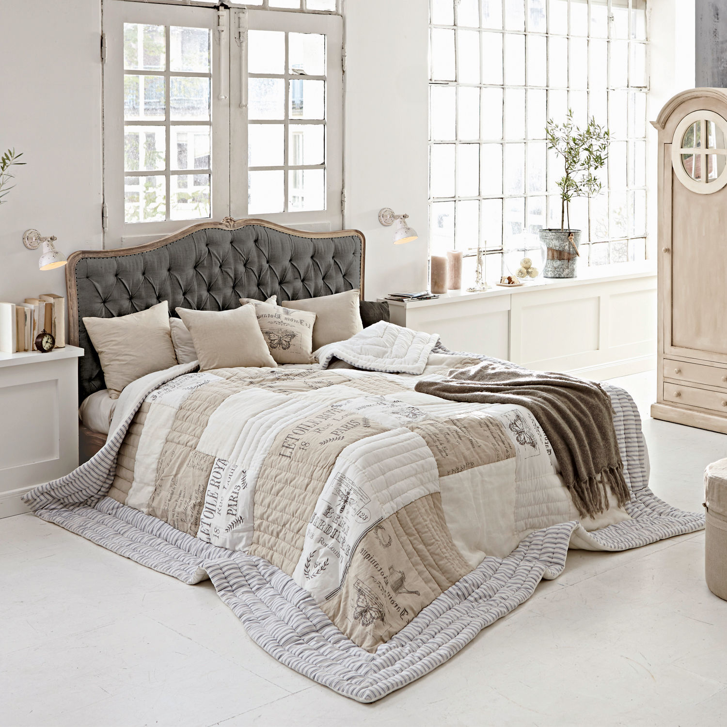 Letto Declaire Camera accogliente con letto con testiera imbottita grigia e copriletto beige e bianco con stampa vintage