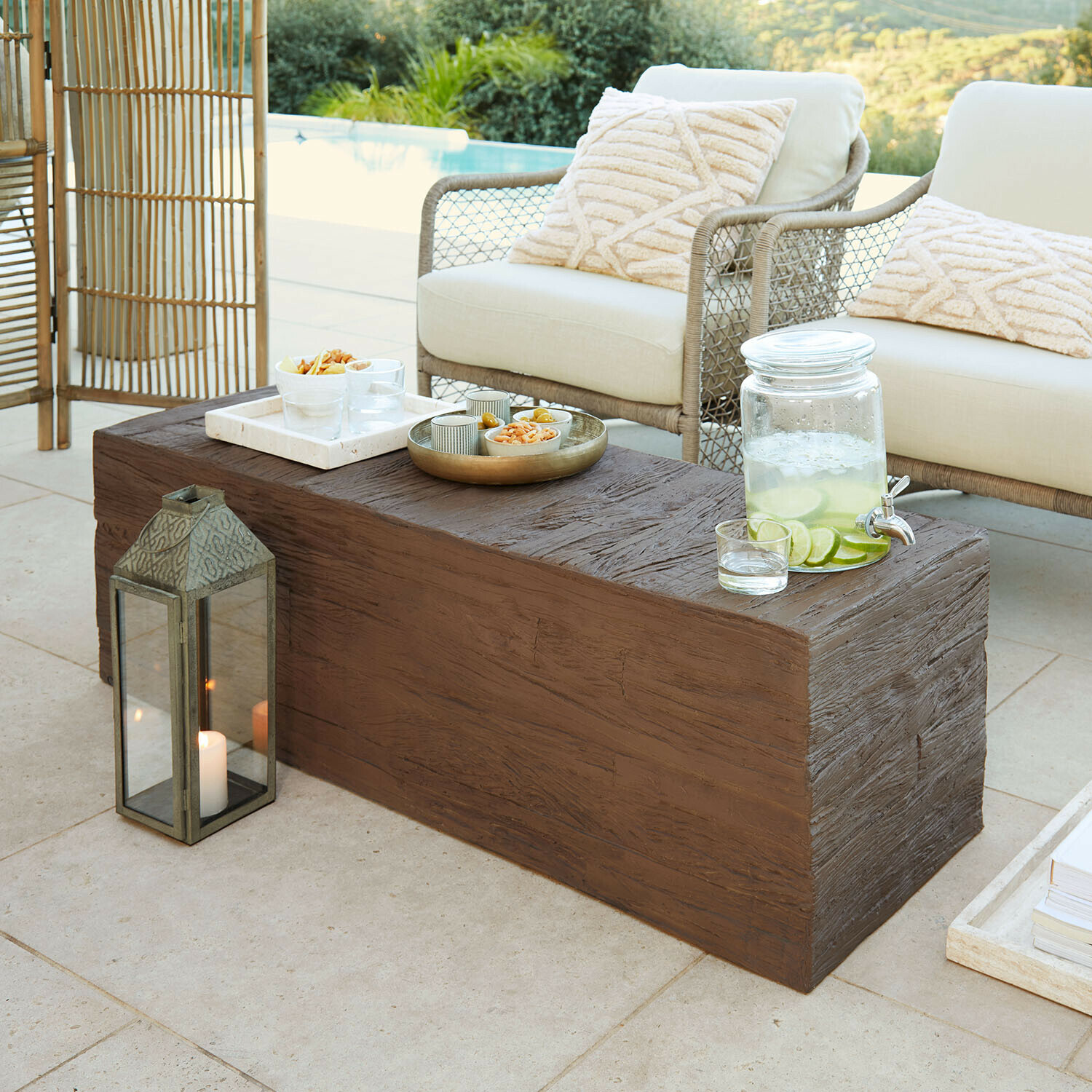 Tavolino marrone effetto legno su terrazza con cuscini crema e brocca di limonata