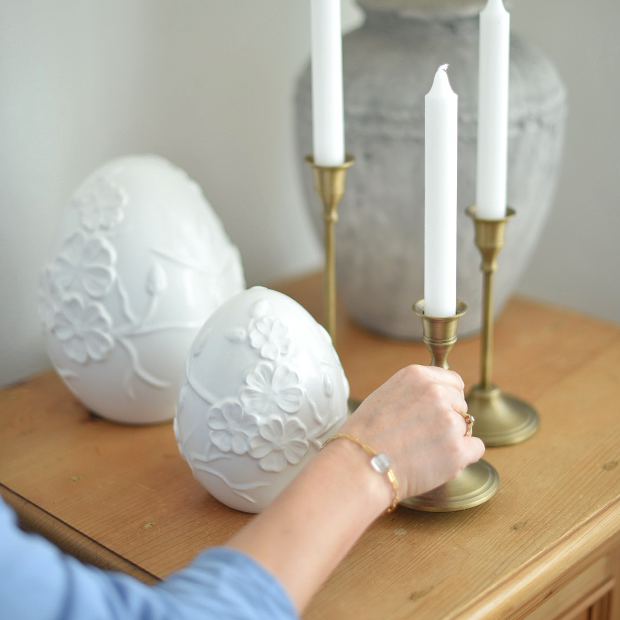 Uova decorative in ceramica bianca con rilievo floreale su tavolo in legno accanto a candelabri in ottone