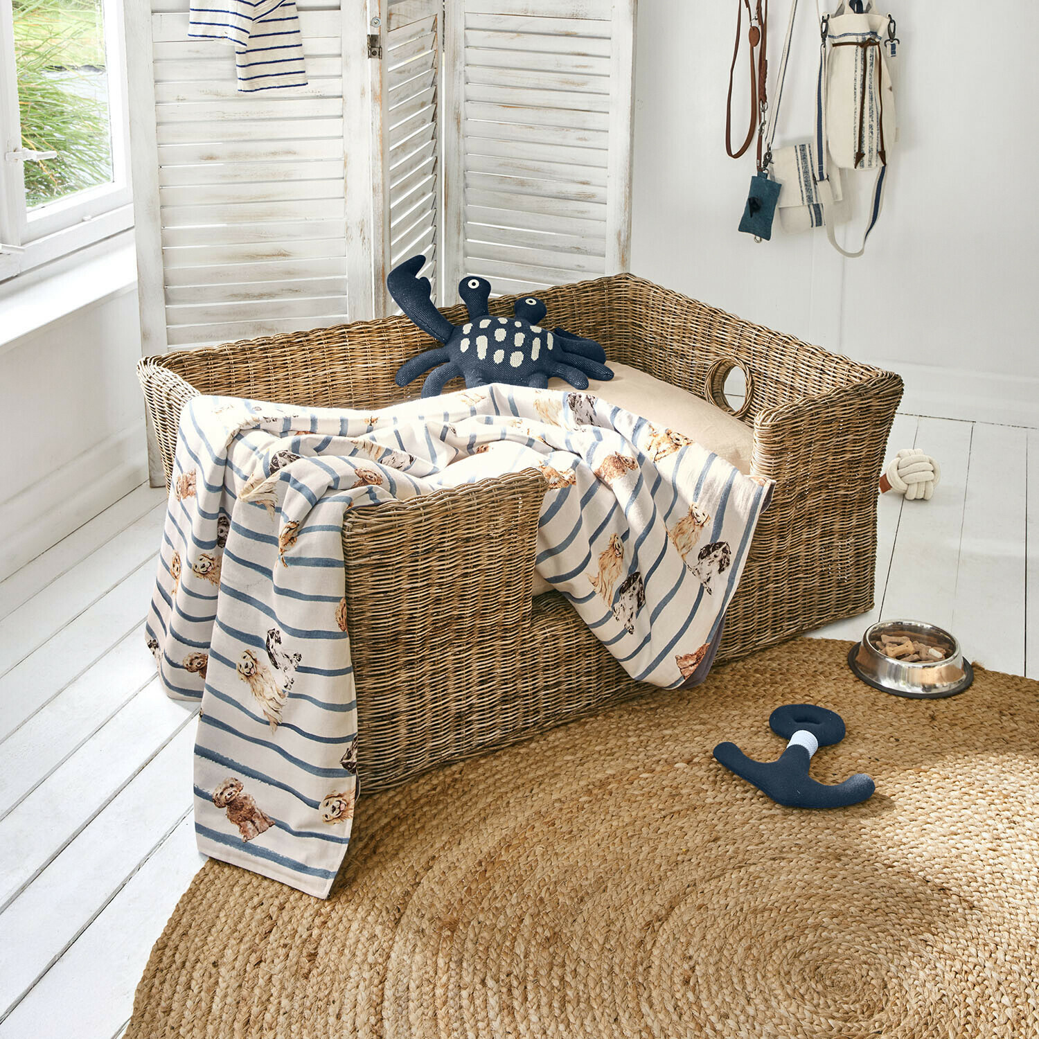 Cuccia per animali Knuddo Cuccia in vimini per cane con cuscino beige e coperta a righe con motivo cani, giocattoli e ciotola sul pavimento