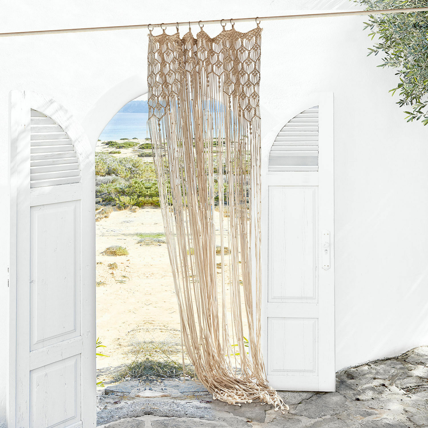 Tenda Manville Macramè beige in cotone appeso a un ingresso bianco aperto con vista sulla spiaggia e sul mare