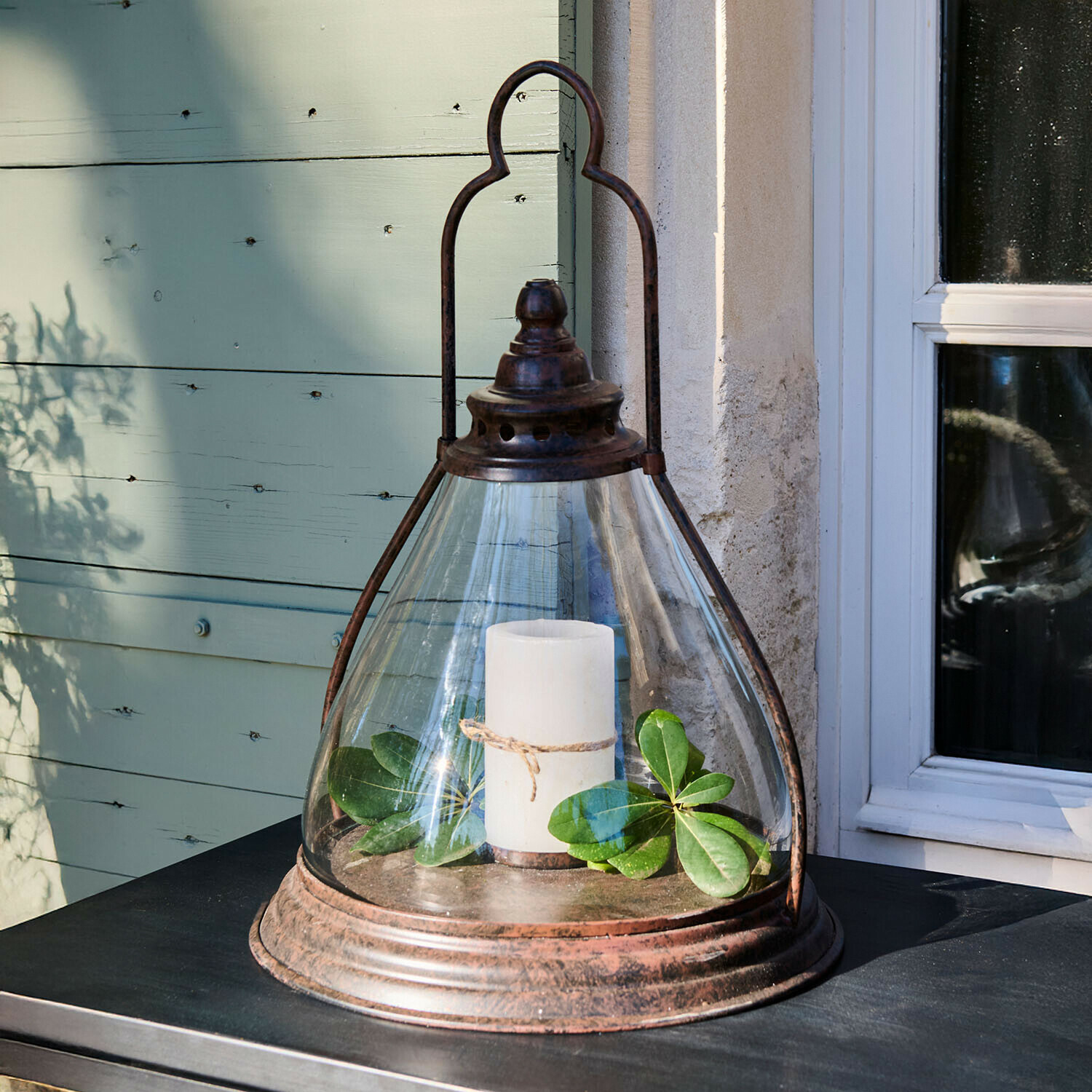 Lanterna in vetro con struttura metallica arrugginita, candela bianca e foglie verdi su tavolo