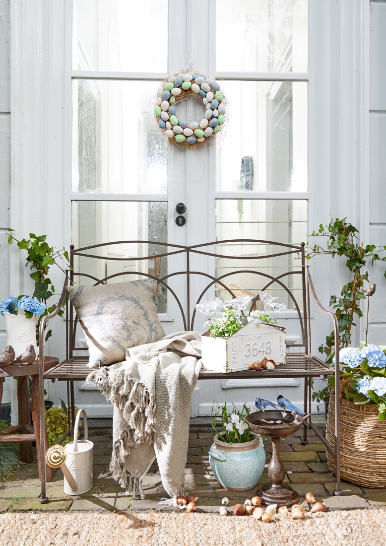 Panchina da giardino in metallo con coperta beige e cuscino, decorazioni floreali e corona di uova pastello