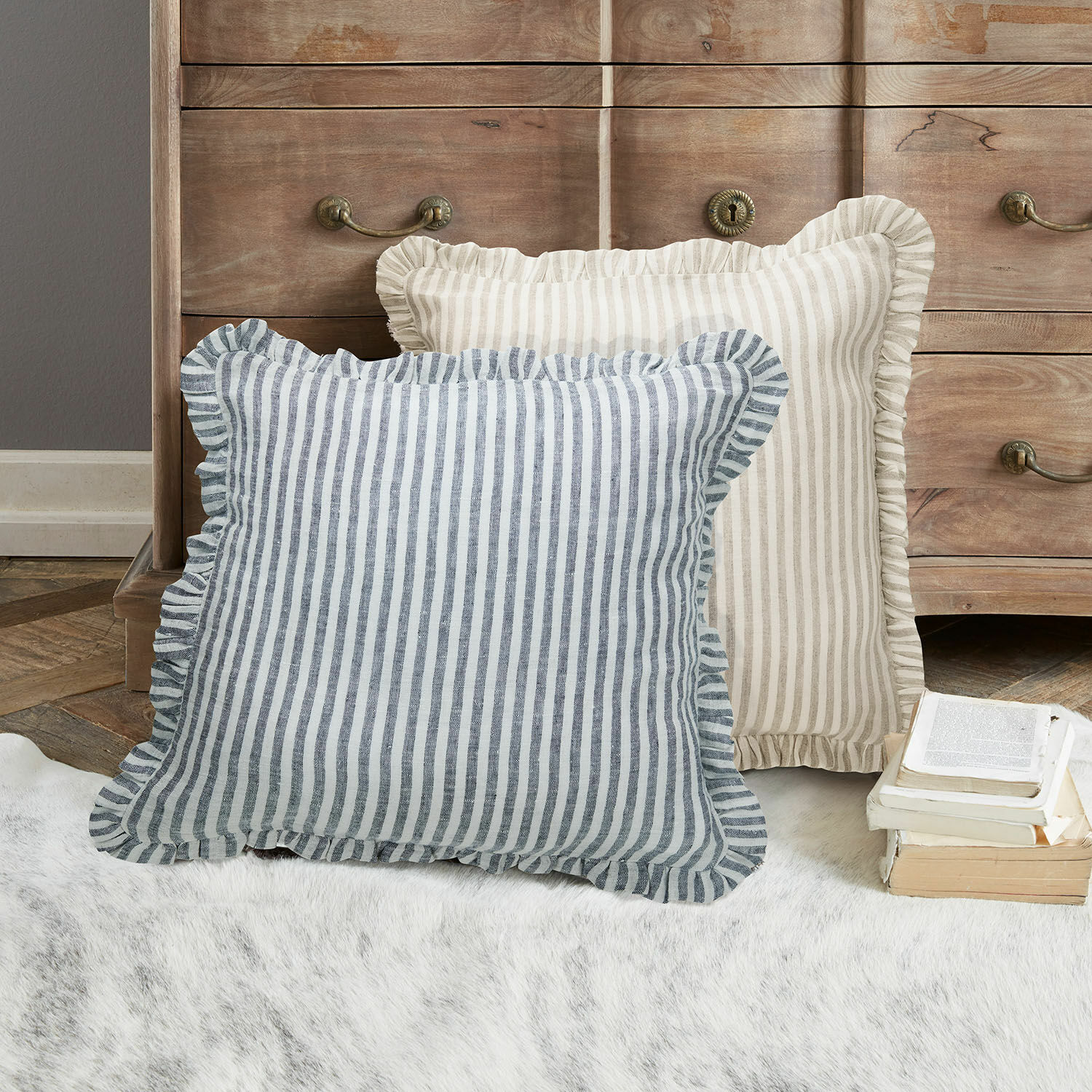 Coppia di copricuscini Brenda Due cuscini decorativi a righe con volant beige e blu su tappeto davanti a una cassettiera in legno