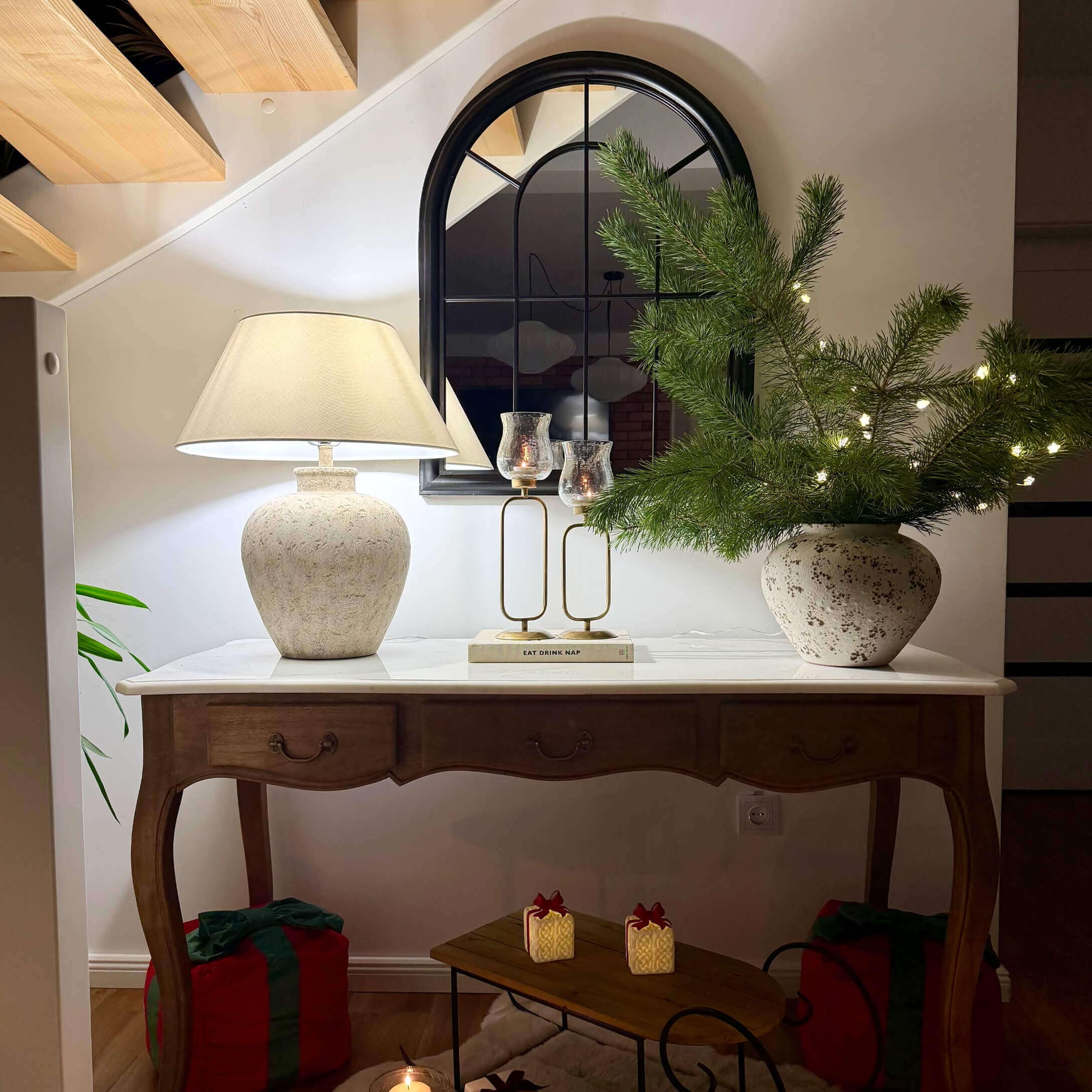 Tavolo in legno con piano bianco, lampada in ceramica, specchio e decorazioni natalizie con rami verdi e luci