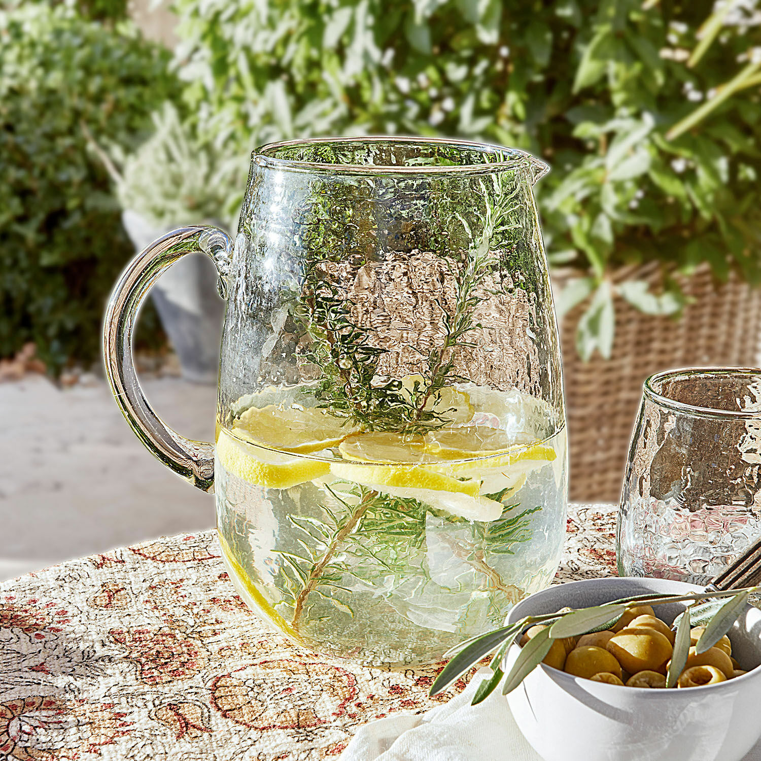 Caraffa Rezay Brocca di vetro con acqua, limone e erbe su tovaglia decorativa, bevanda estiva in terrazza