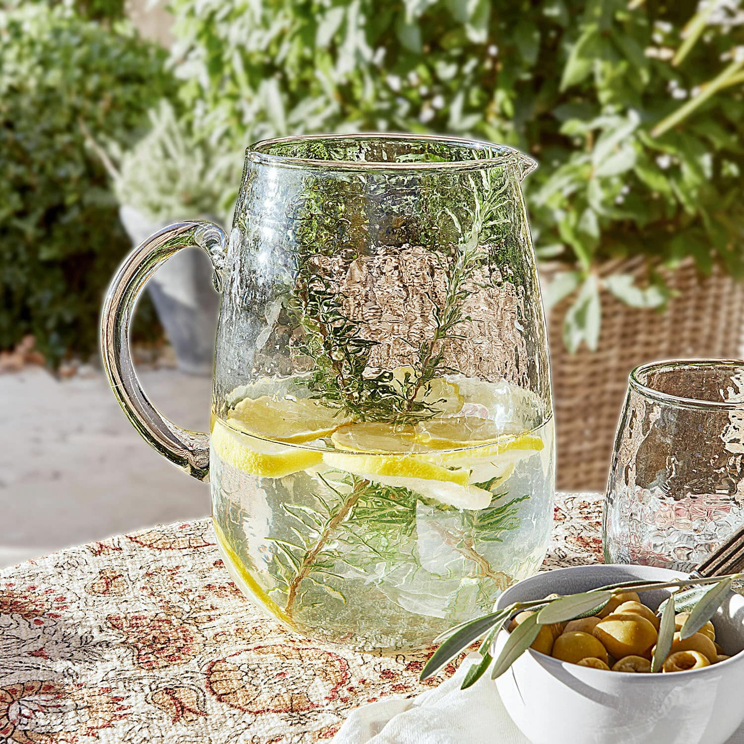Caraffa Rezay Brocca di vetro con acqua, limone e erbe su tovaglia decorativa, bevanda estiva in terrazza