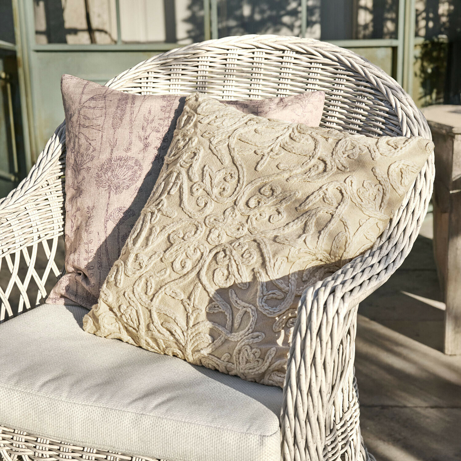 Coppia di copricuscini Floraine Due cuscini decorativi beige e rosa su una poltrona in vimini bianco su un terrazzo soleggiato