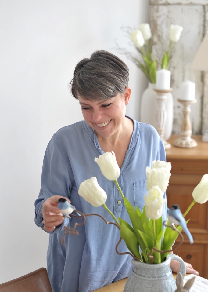 Donna con camicia azzurra con tulipani bianchi e uccelli decorativi in brocca grigia sul tavolo