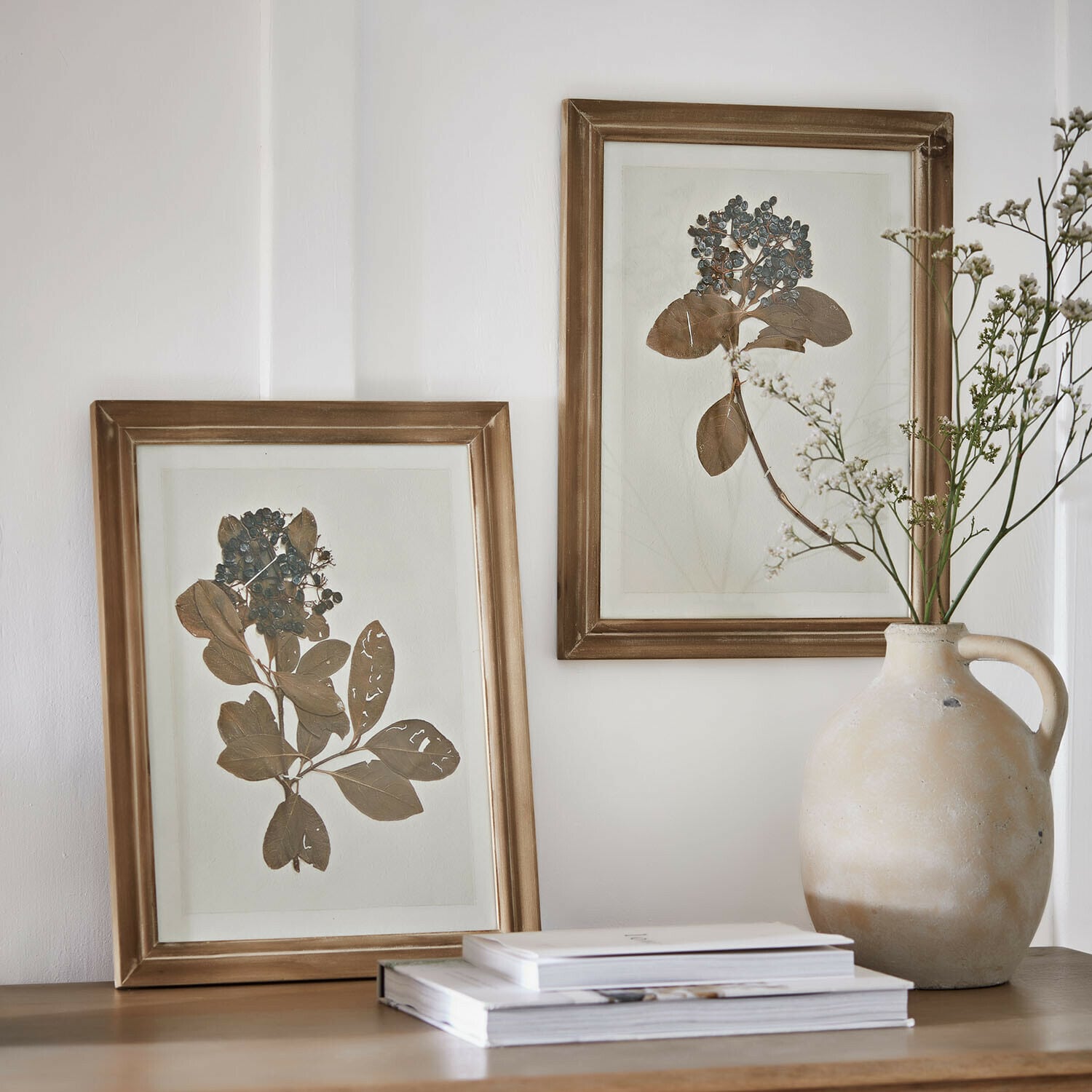 Due cornici dorate con foglie e bacche essiccate su tavolo in legno accanto a vaso in ceramica e libri