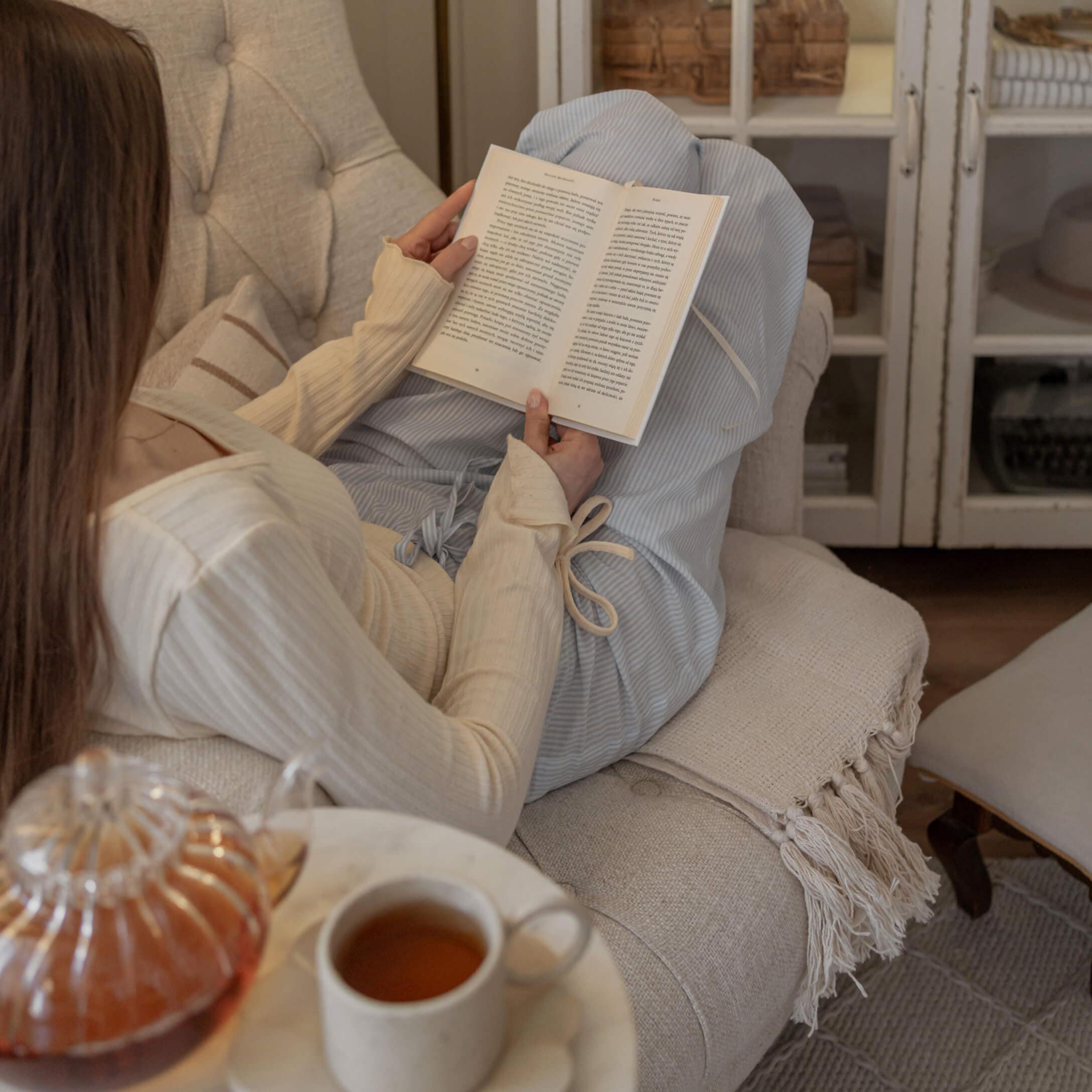 Donna con maglione crema e pantaloni blu legge un libro su divano beige con tazza di tè sul tavolo