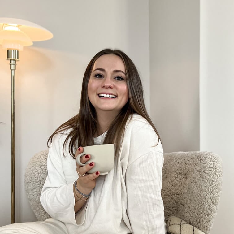 Donna sorridente con maglione bianco che tiene una tazza beige seduta su poltrona morbida in ambiente luminoso