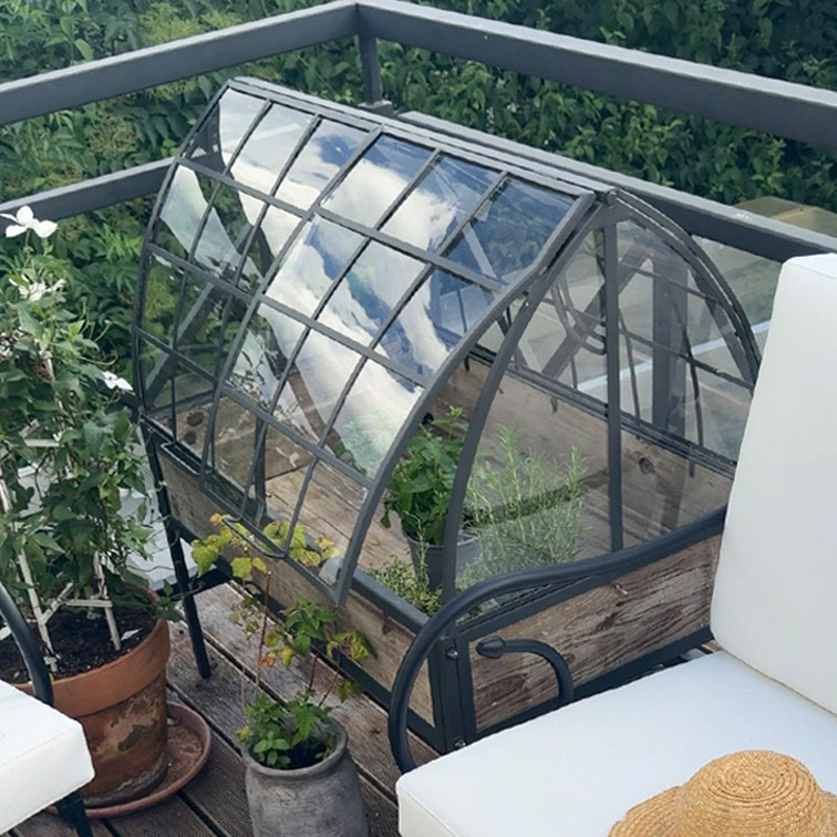 Piccola serra da giardino con struttura in metallo e base in legno su terrazza con piante e mobili da esterno