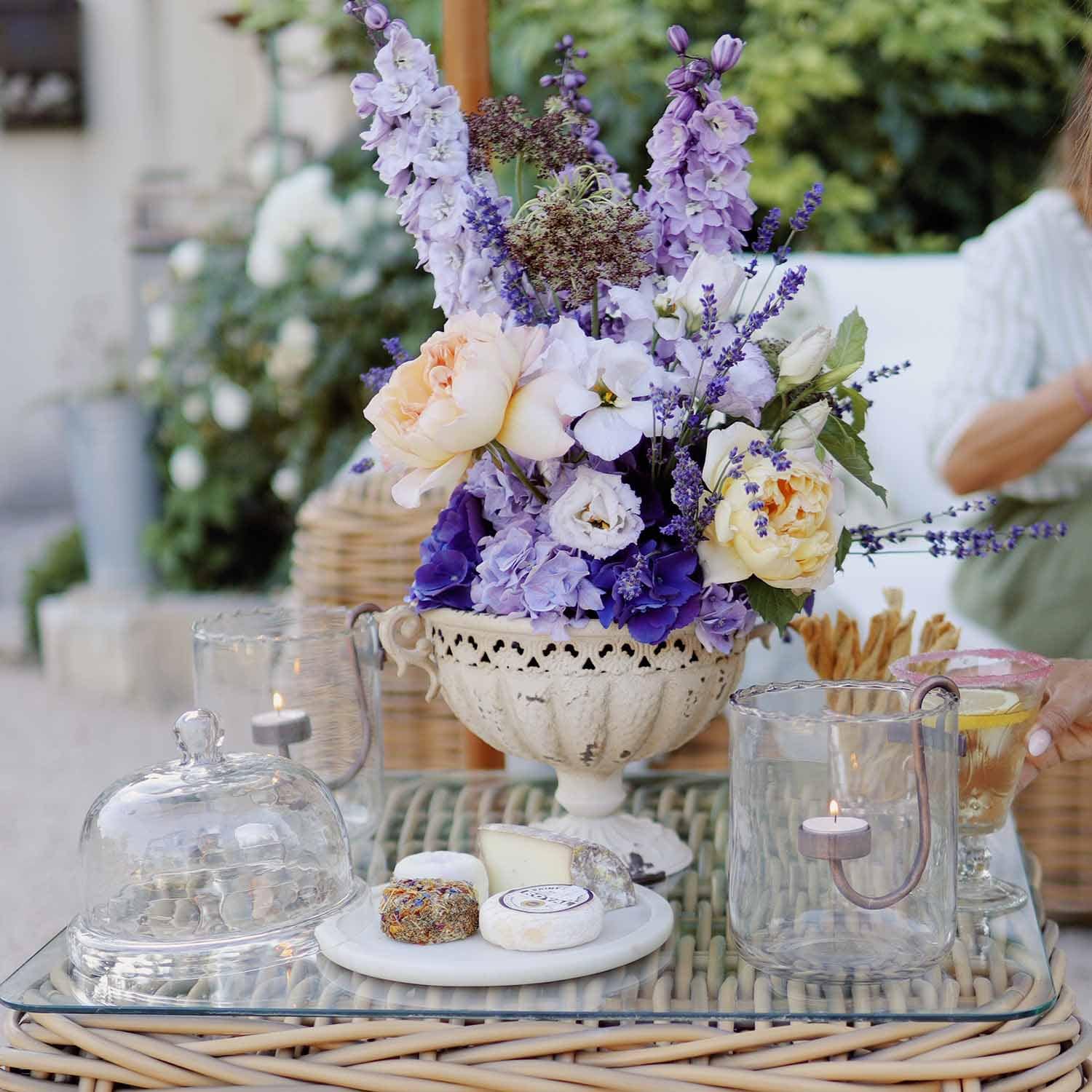 Composizione di fiori viola e crema in vaso di ceramica bianca su tavolo di vetro con candele e formaggi